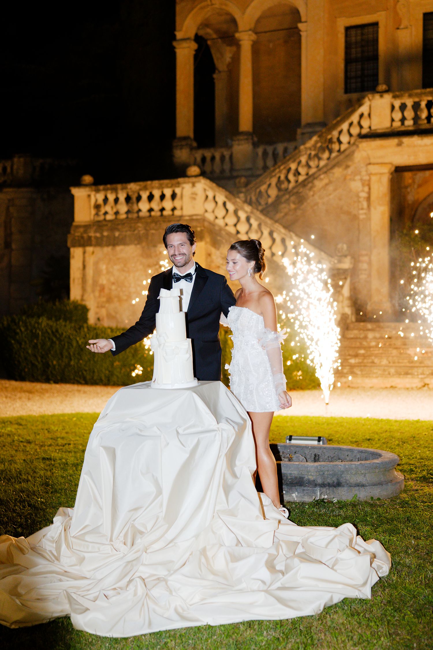 A couple cuts their wedding cake outdoors with sparklers and a grand staircase in the background.