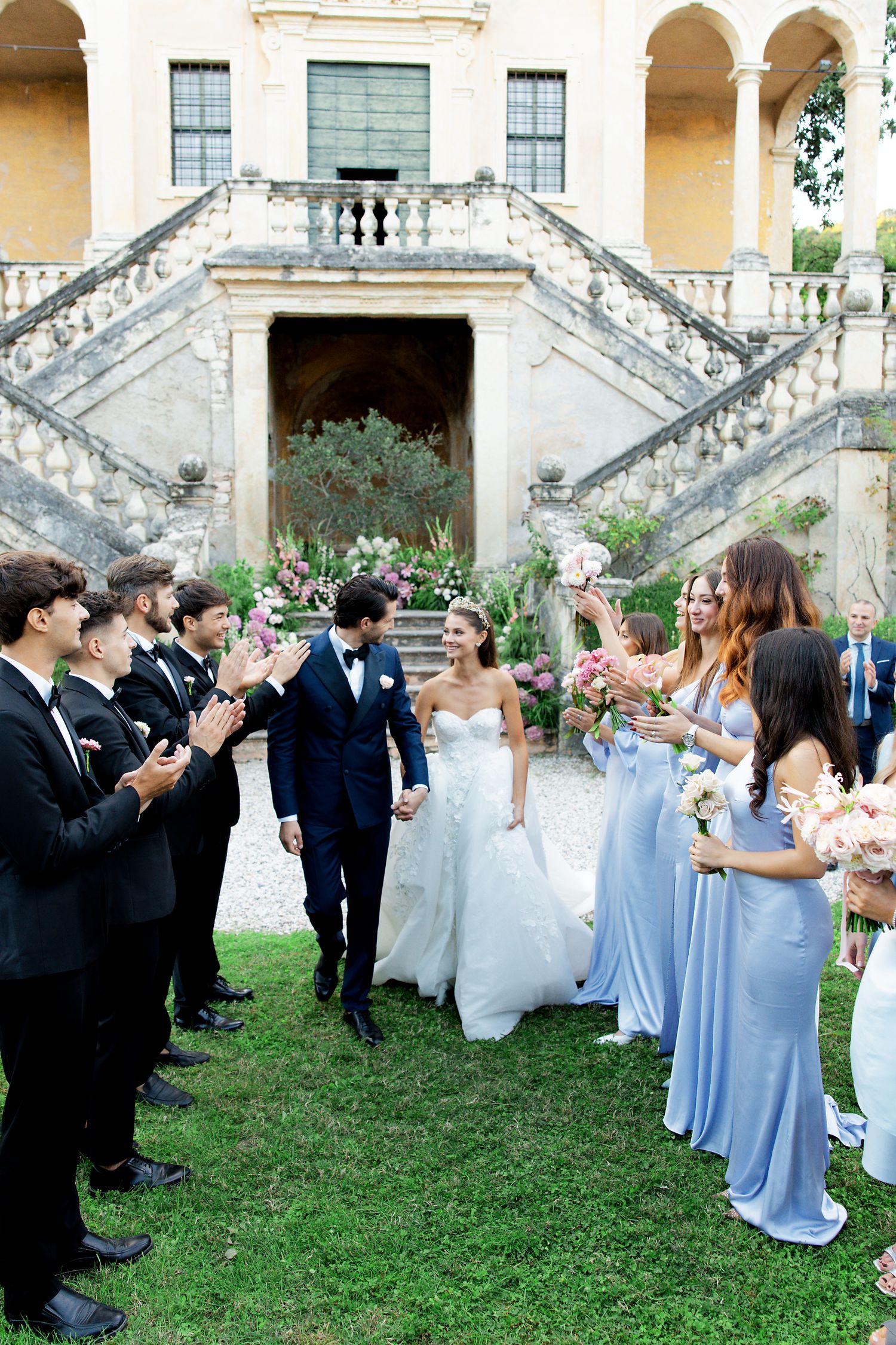 A wedding party celebrates outside a grand villa with bridesmaids in light blue dresses and groomsmen in black suits.