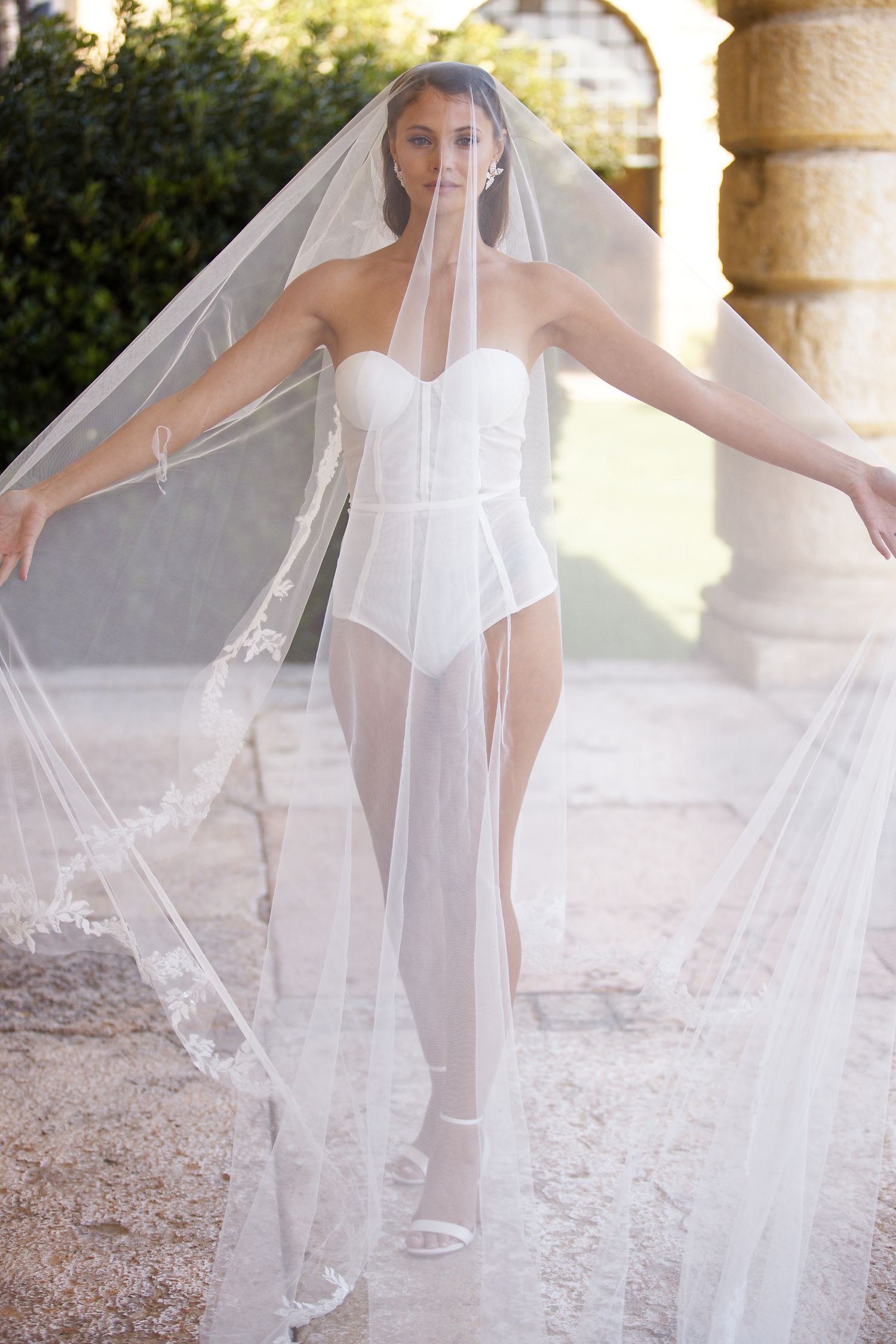 Ethereal bridal portrait with flowing veil and white bodysuit in outdoor garden setting.