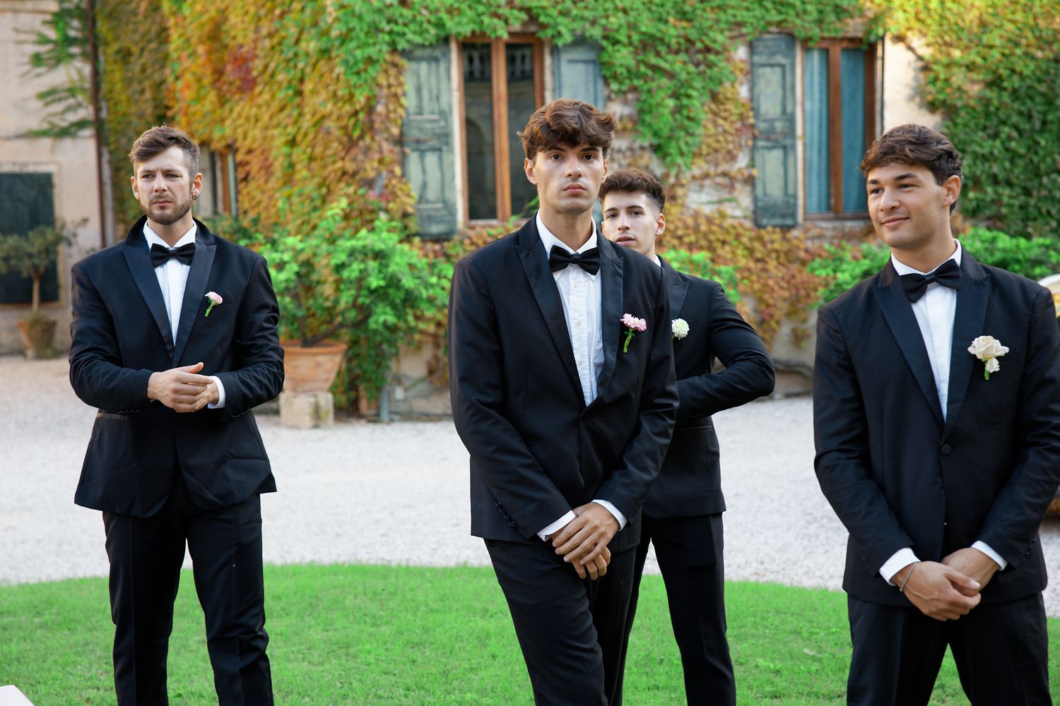 Groomsmen in black tuxedos and bow ties standing on lawn in front of ivy-covered building.