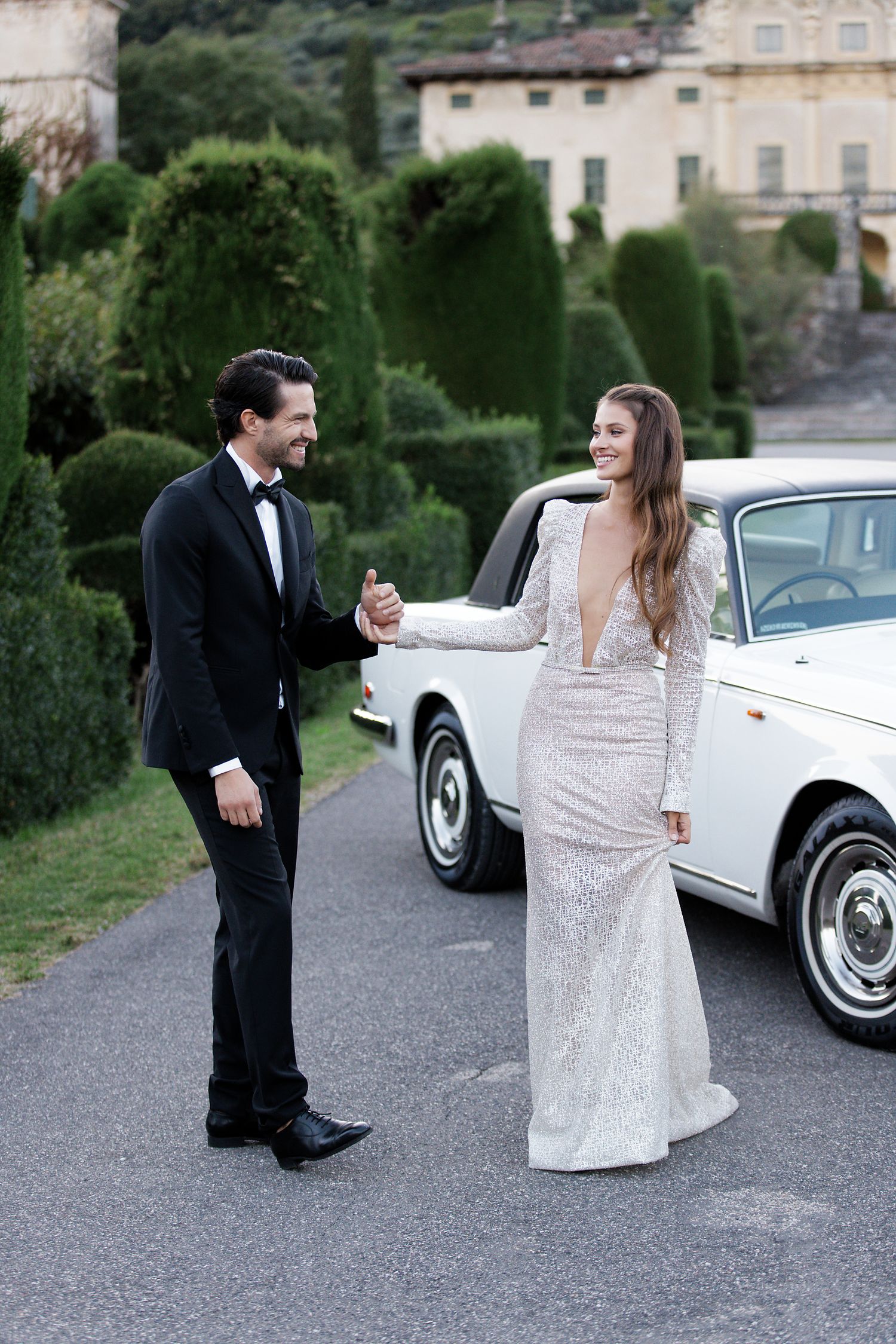 A couple in formal attire share a playful moment on a driveway with a vintage white car.