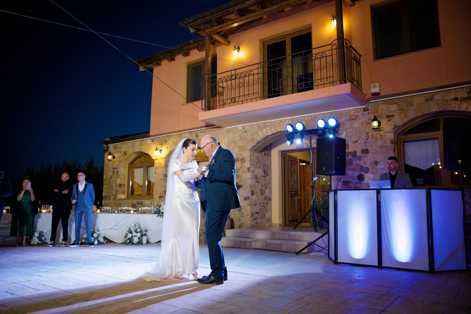 Evening wedding dance under romantic lighting at a Mediterranean-style venue with stone walls.