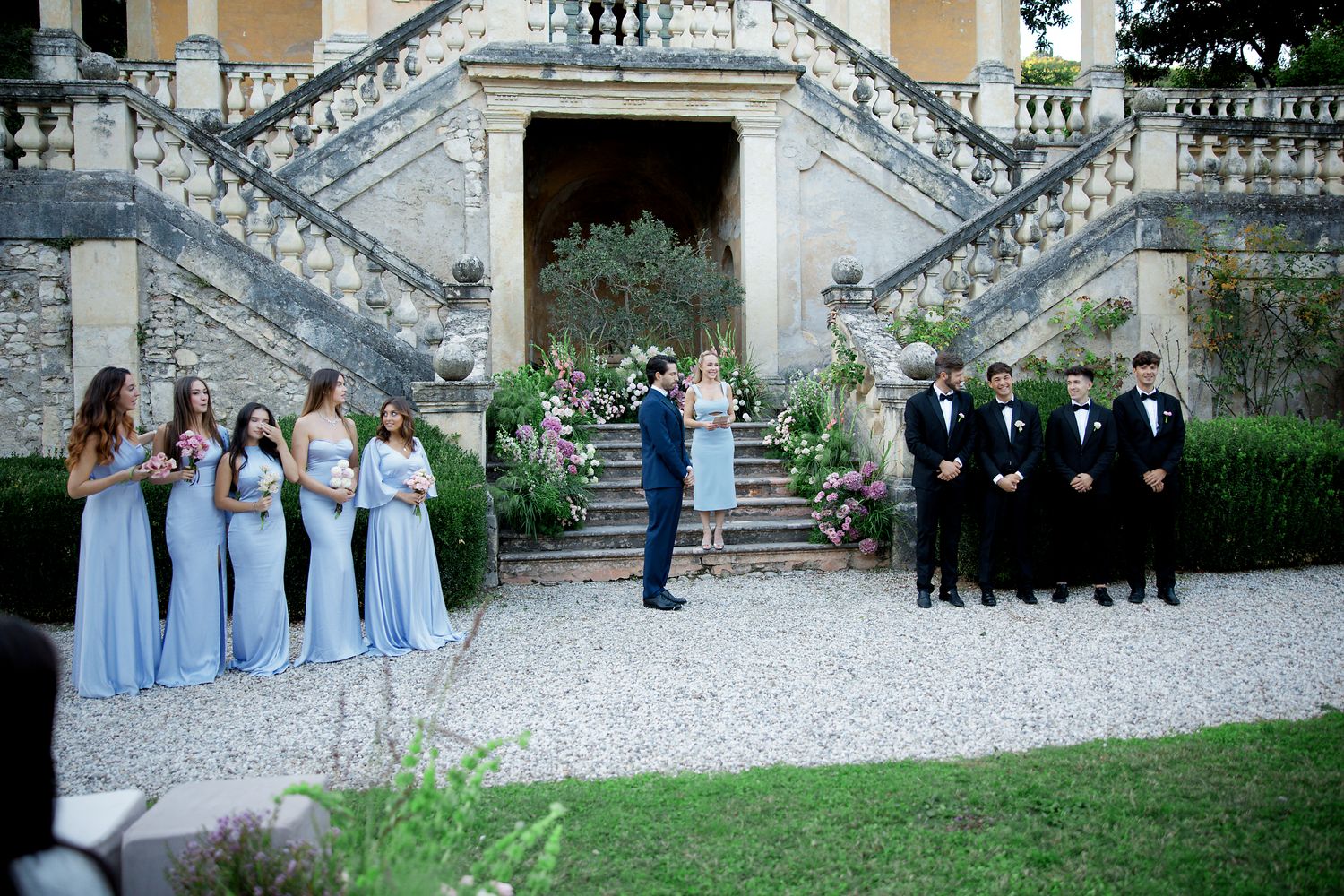 Wedding party in light blue dresses and black suits posed on grand stone staircase.