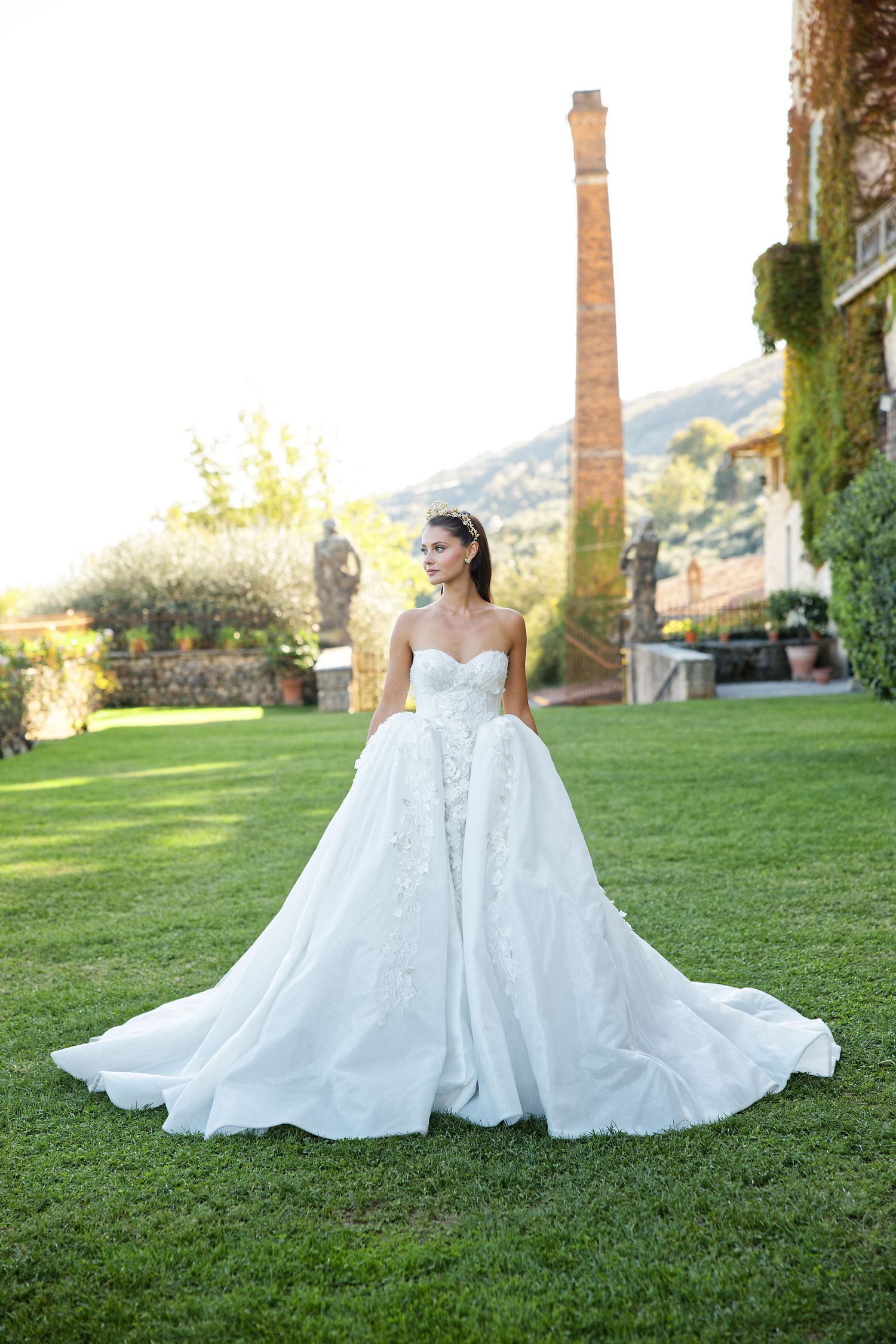 Elegant white wedding gown with full skirt and train photographed on manicured lawn with brick chimney in background.