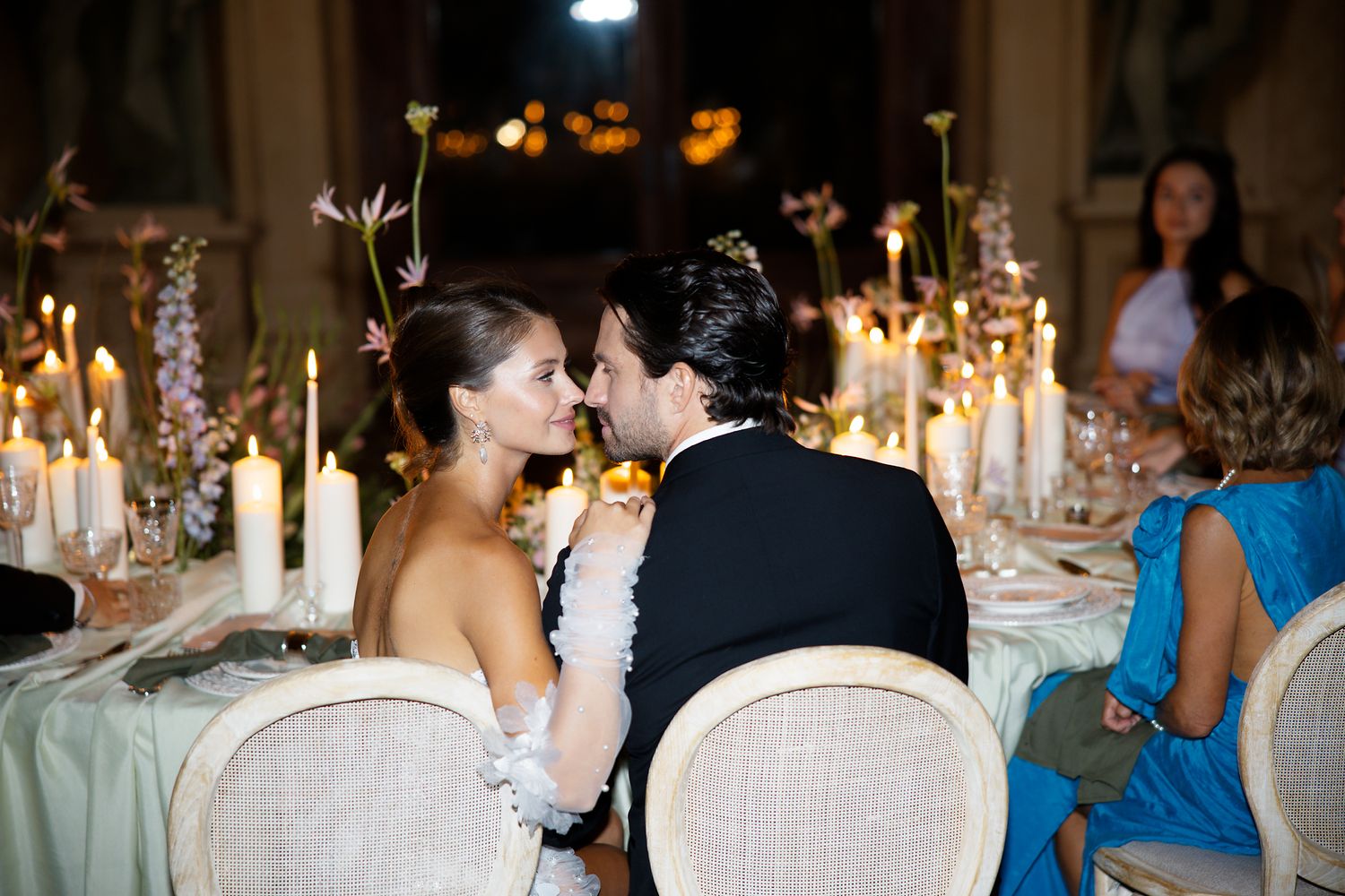 A romantic candlelit reception dinner with guests seated at an elegantly decorated table with tapered candles and floral arrangements.