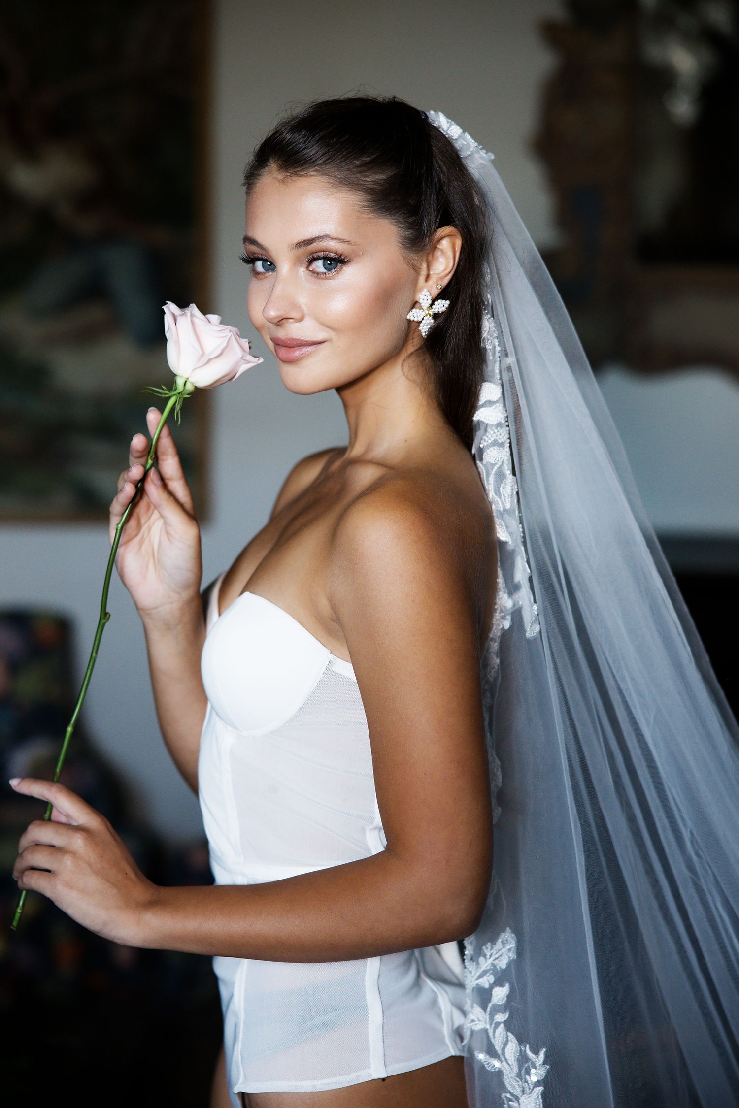 Bride in a white corset holding a single rose while wearing an elegant lace-trimmed veil.