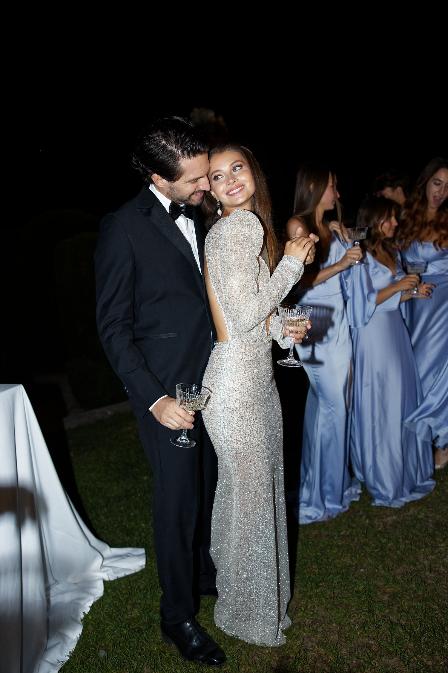 Elegant couple in formal wear enjoying drinks at an outdoor evening celebration with guests in blue dresses.