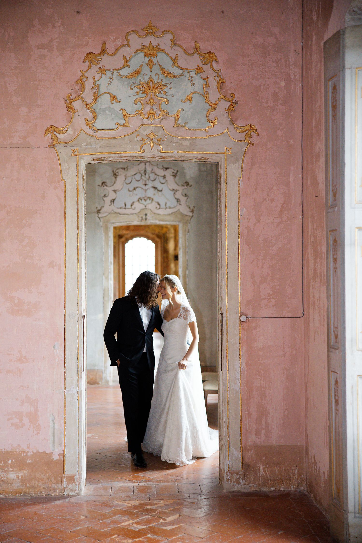 A couple shares an intimate moment in an ornate doorway with baroque architectural details and pink walls.