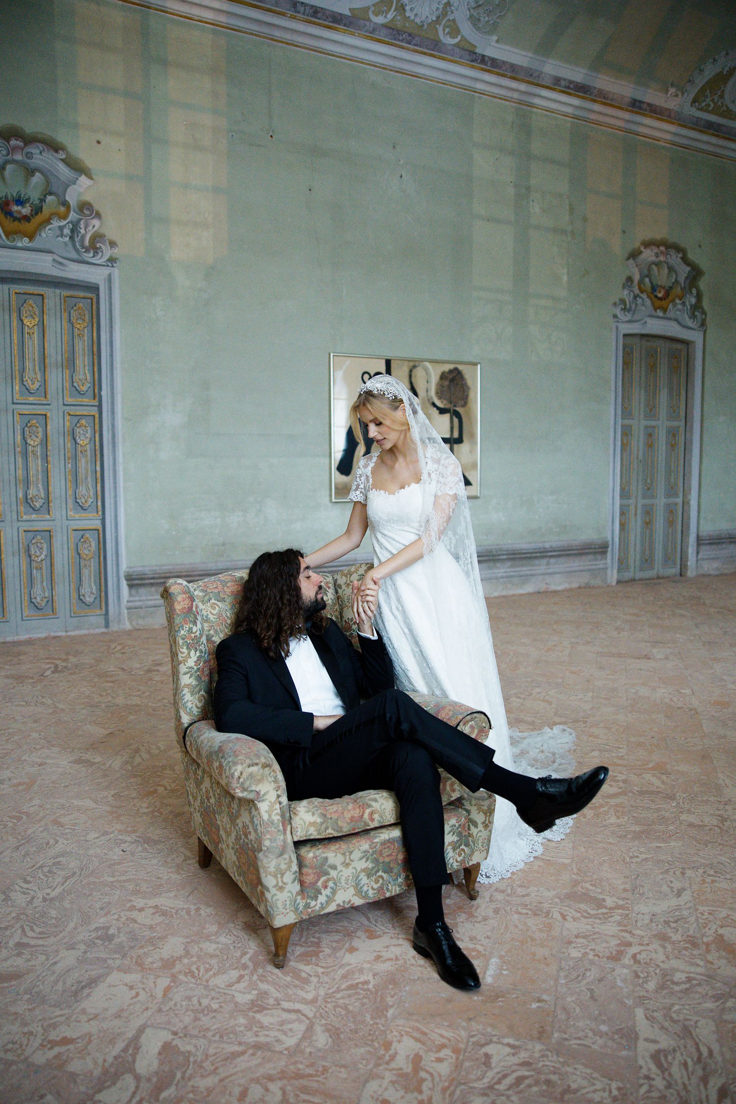 Two individuals in formal wedding attire share a tender moment in an elegant room with ornate doors and decor.