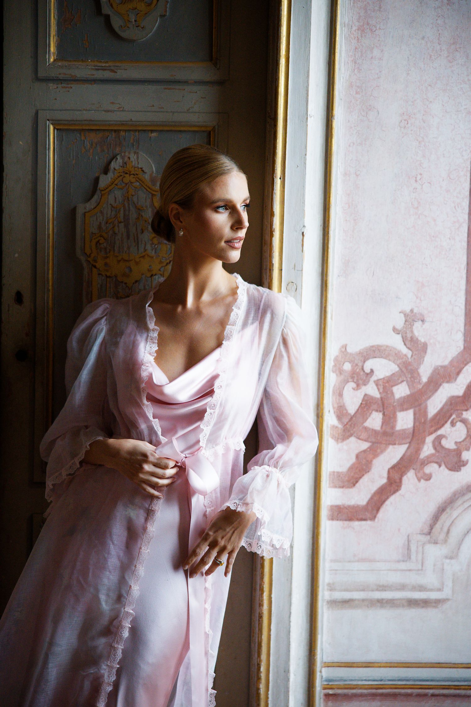 A delicate pink silk robe catches the light near an ornately decorated window in a classical interior.