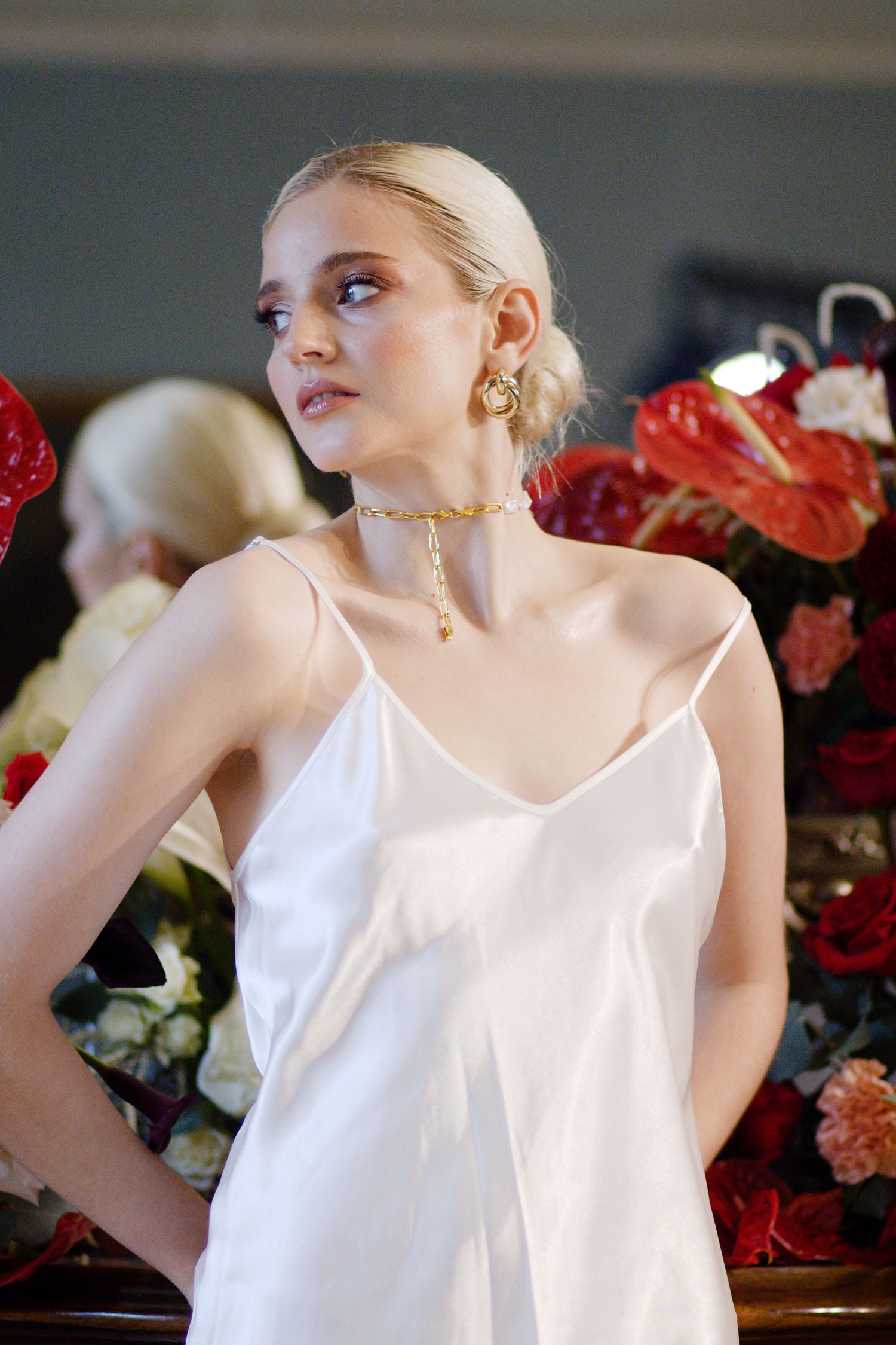 Elegant model in white silk slip dress poses with red floral arrangements and sparkling chandelier.