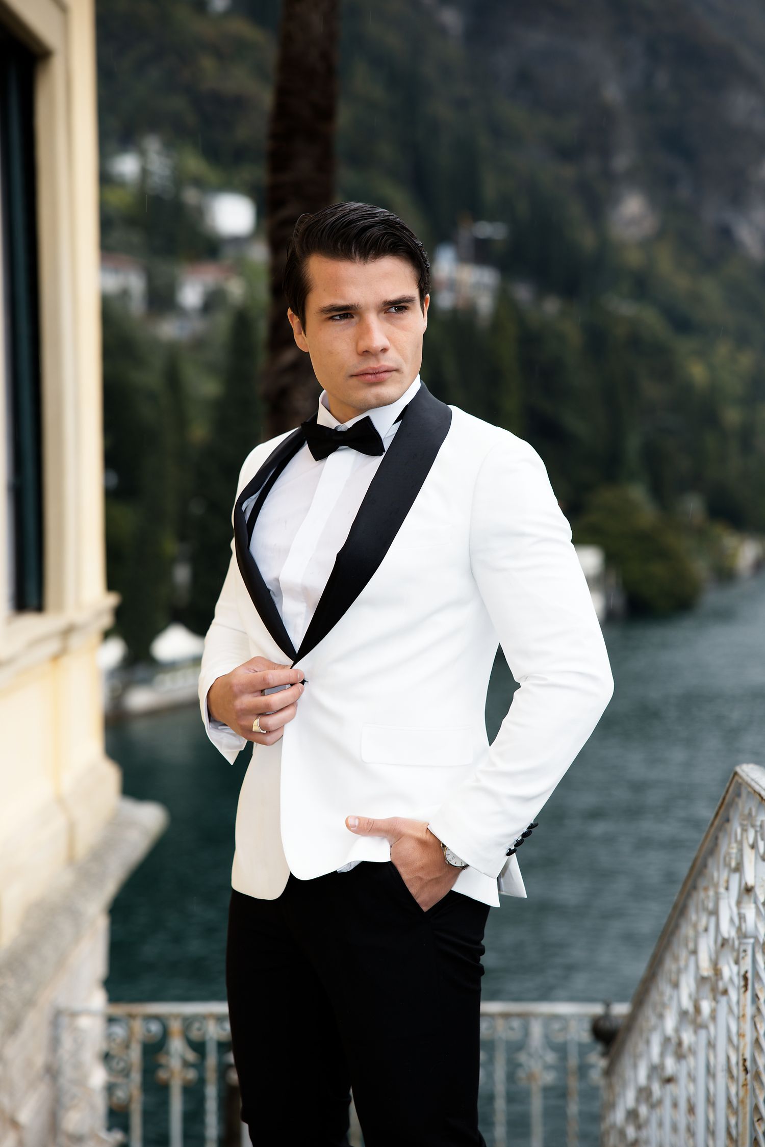 Elegant groom wearing white tuxedo jacket with black lapels poses on balcony overlooking Lake Como.