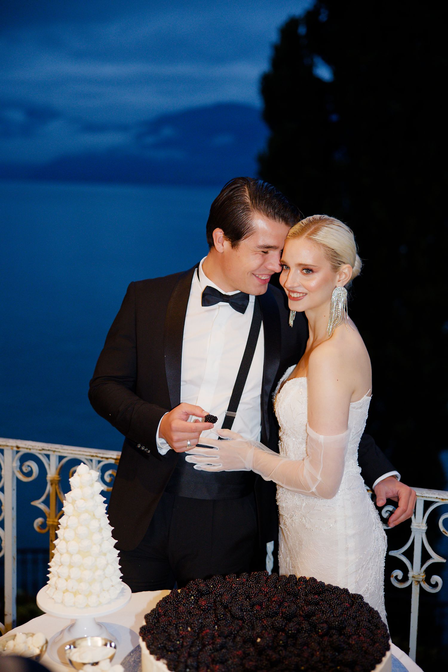 Elegant couple in formal attire share an intimate moment on a balcony with wedding cakes at night.