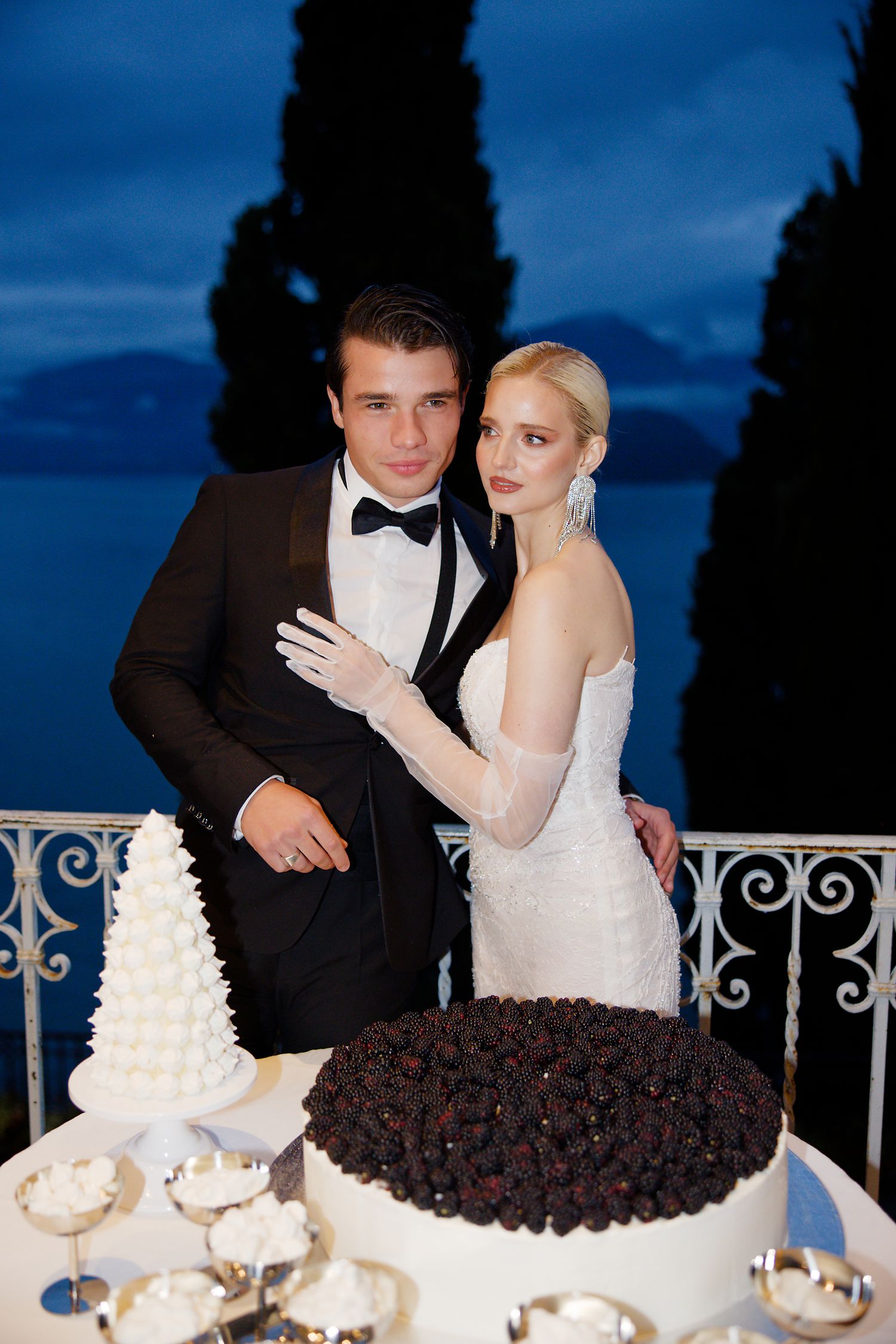 Elegant wedding couple poses by their two-cake display during blue hour on a scenic balcony.