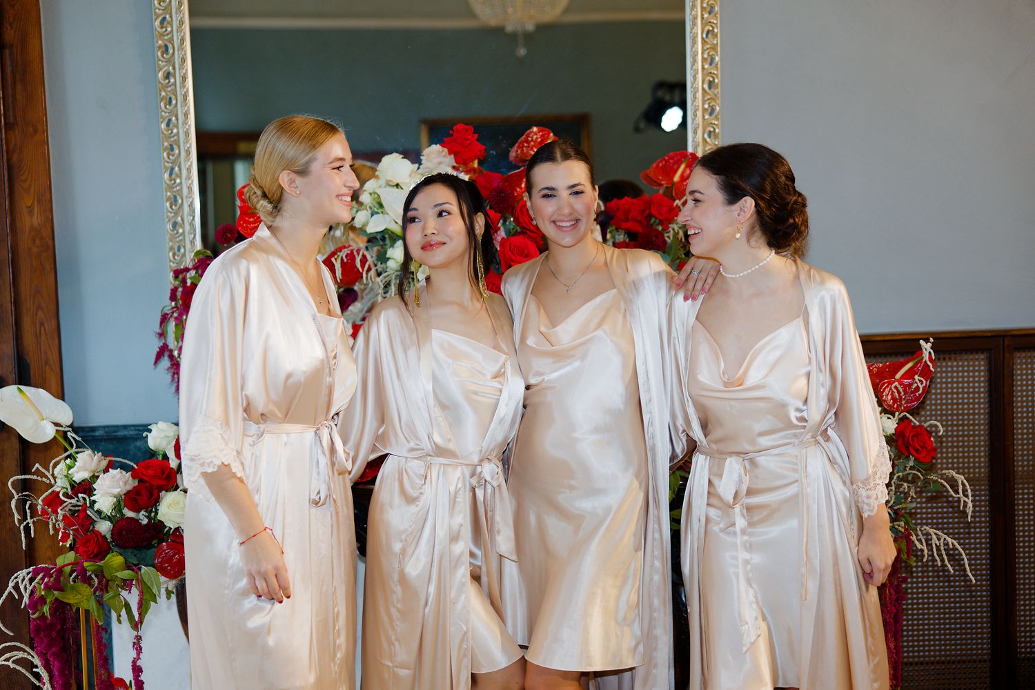Group in matching silk robes share joyful moment with red floral decorations in background.