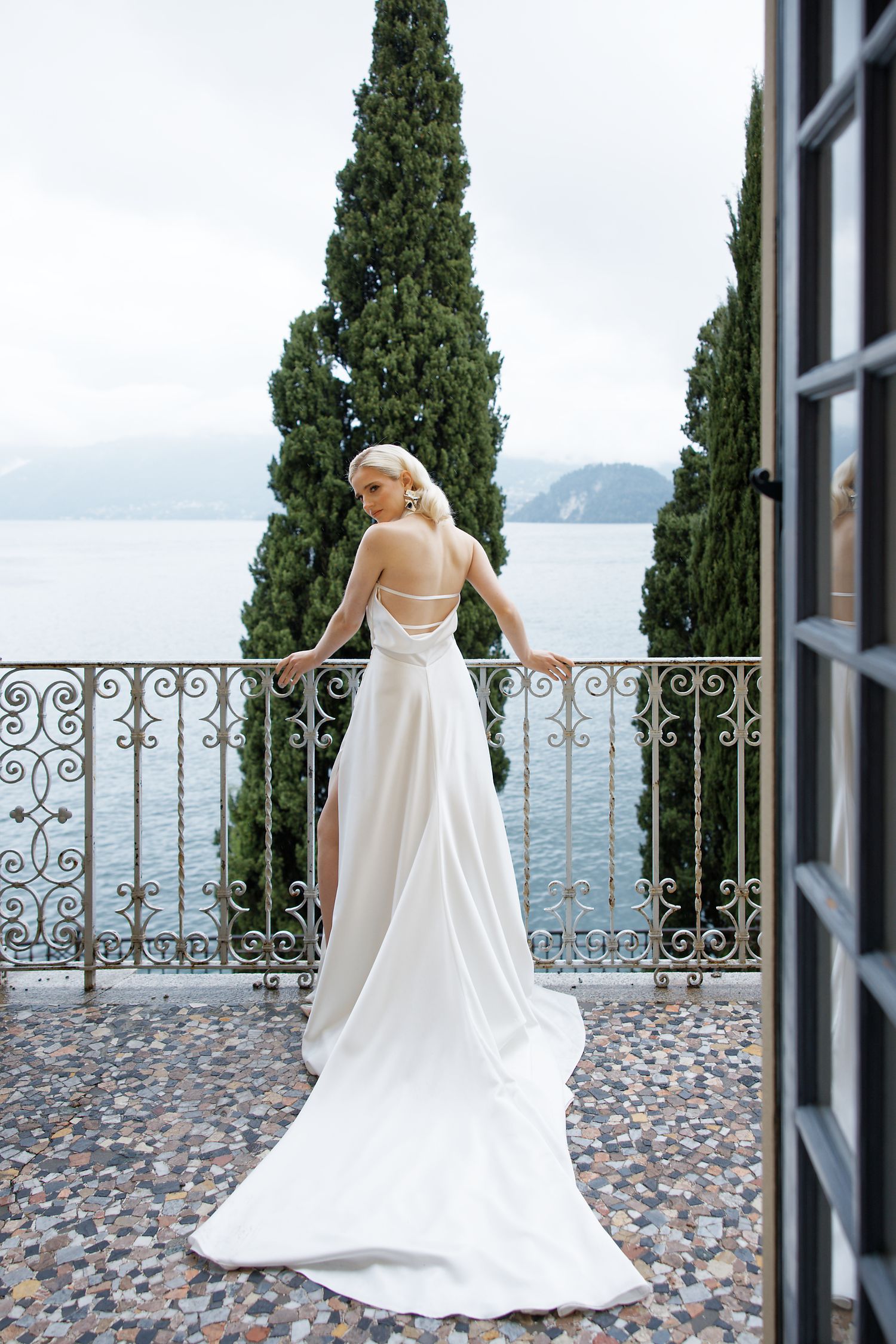 Bride in flowing white gown poses on Mediterranean balcony overlooking serene lake view.