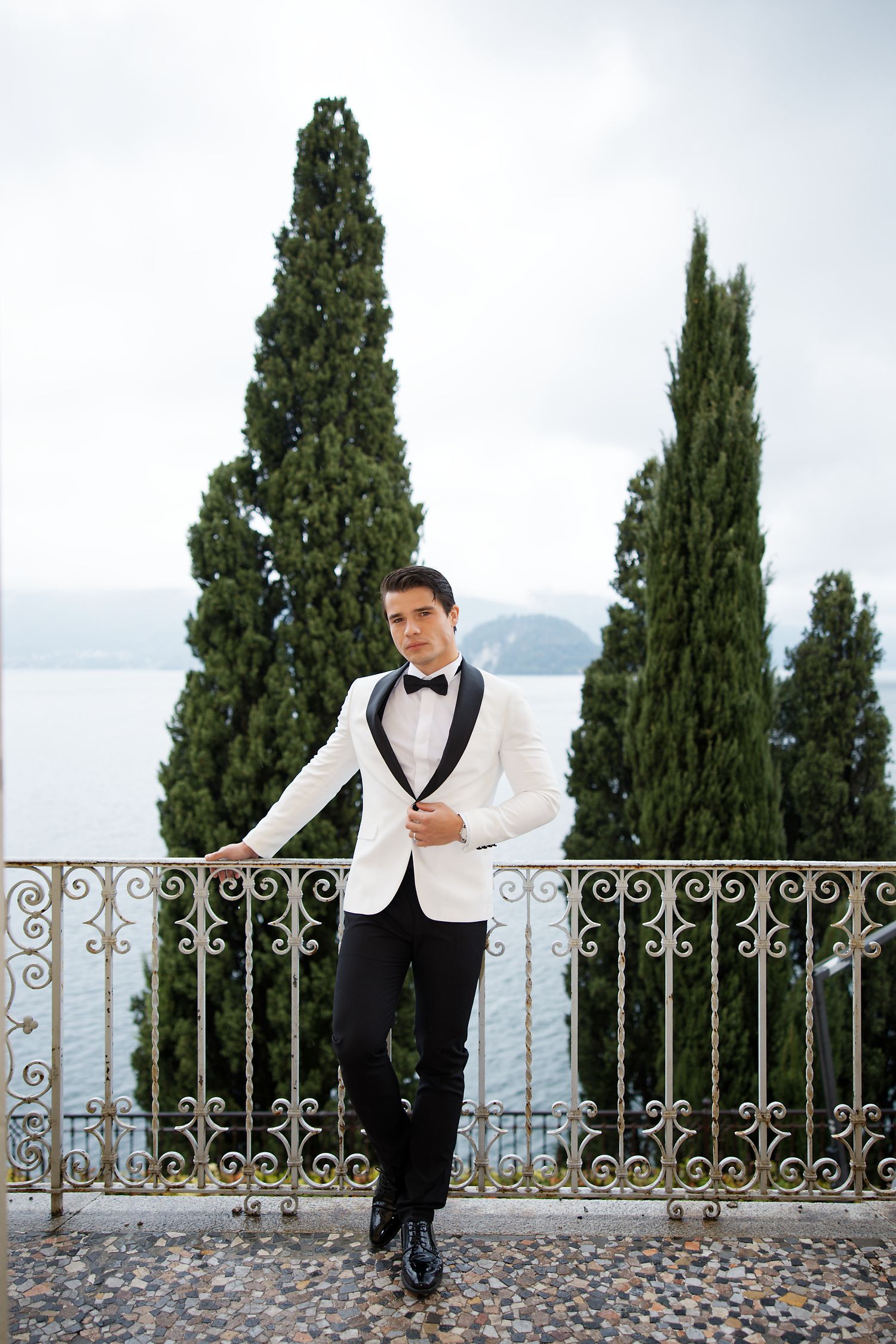Groom in white tuxedo jacket poses against Italian cypress trees and lake view.