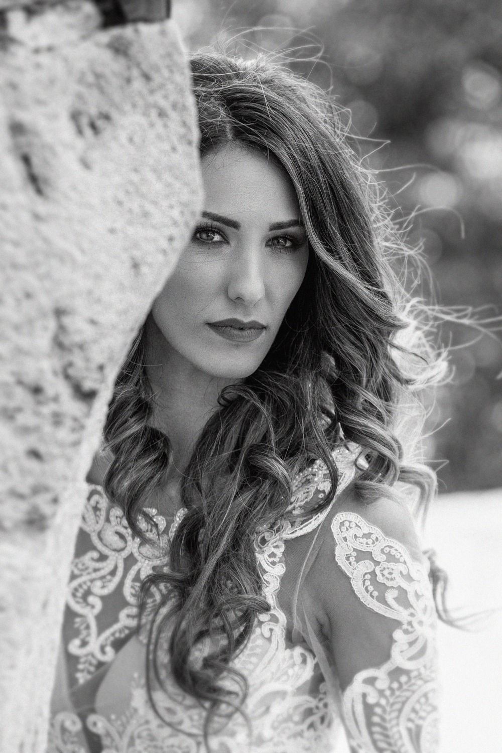 Black and white portrait of someone with long wavy hair wearing a white lace dress leaning against stone wall.