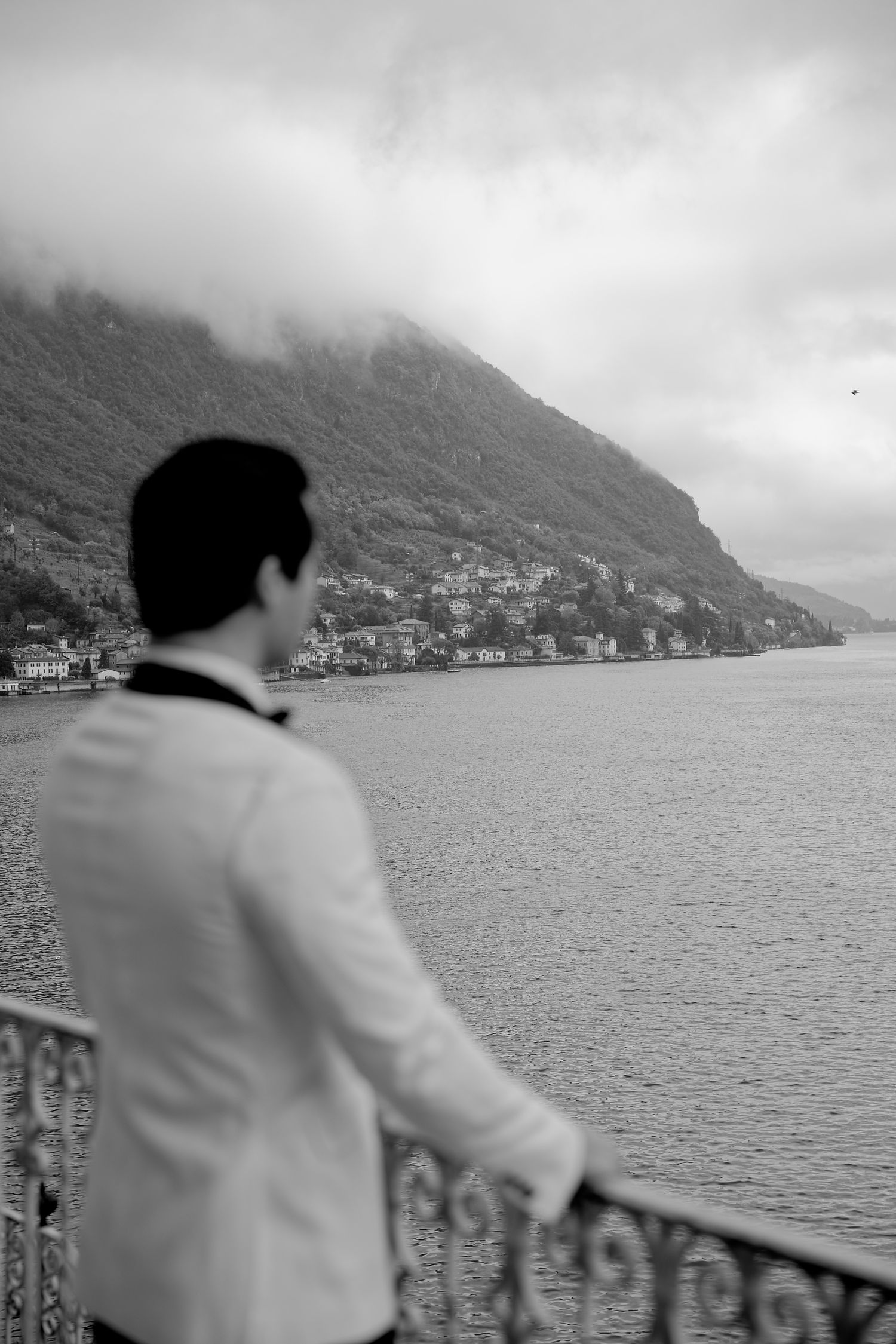 A person in white attire gazes at a misty mountain lake view from an ornate balcony railing in black and white.