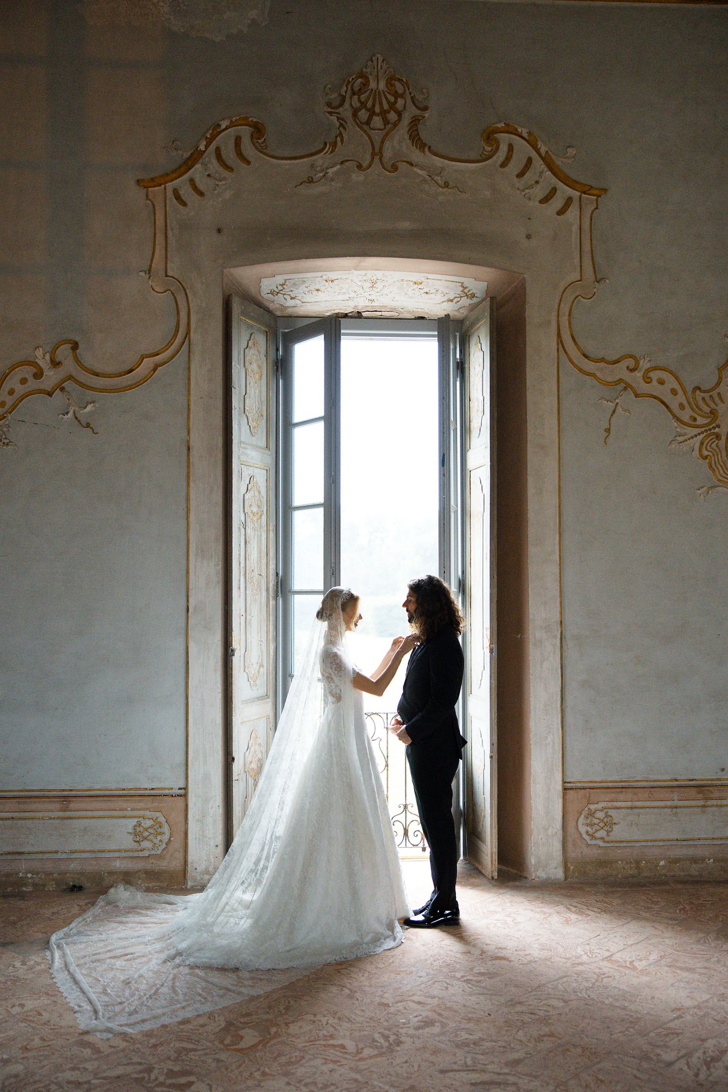 A couple in wedding attire share an intimate moment in a sunlit doorway of an elegant historic building.