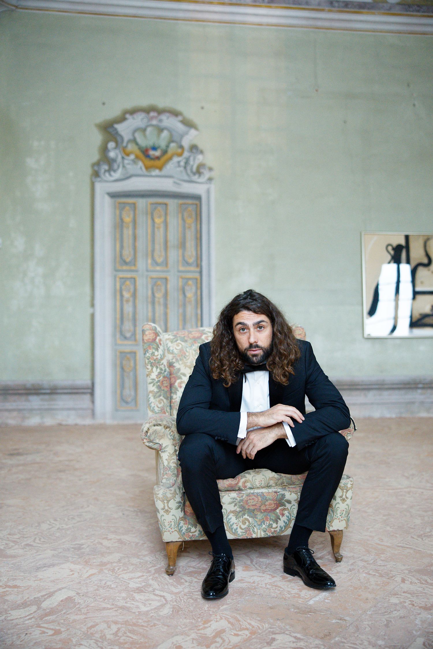Long-haired person in formal black suit sits contemplatively in an ornate vintage armchair against mint green wall.