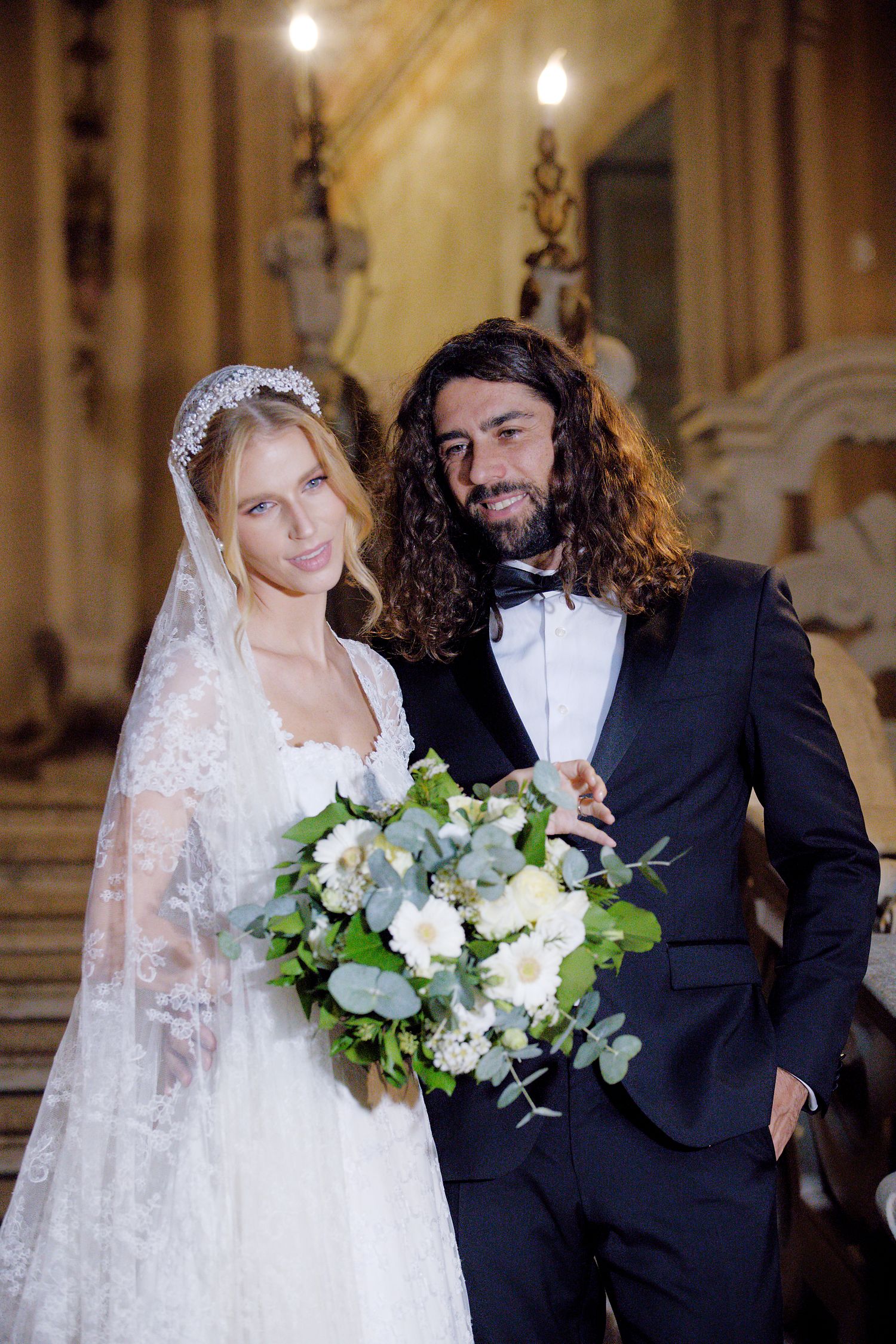 Wedding portrait with lace-veiled bride holding white floral bouquet in elegant church interior.