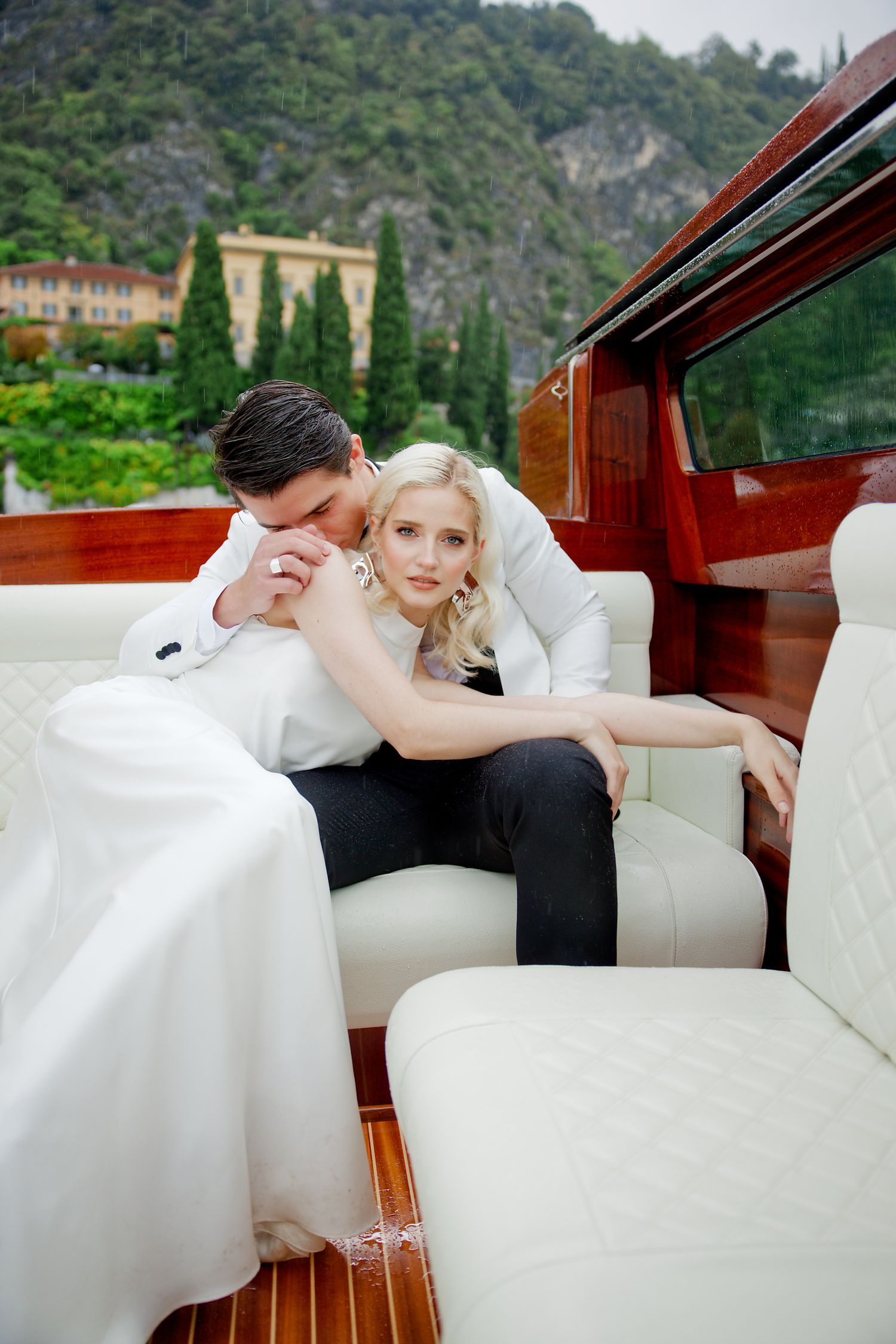 Wedding couple lounging in luxurious boat against backdrop of Italian villa and mountains.