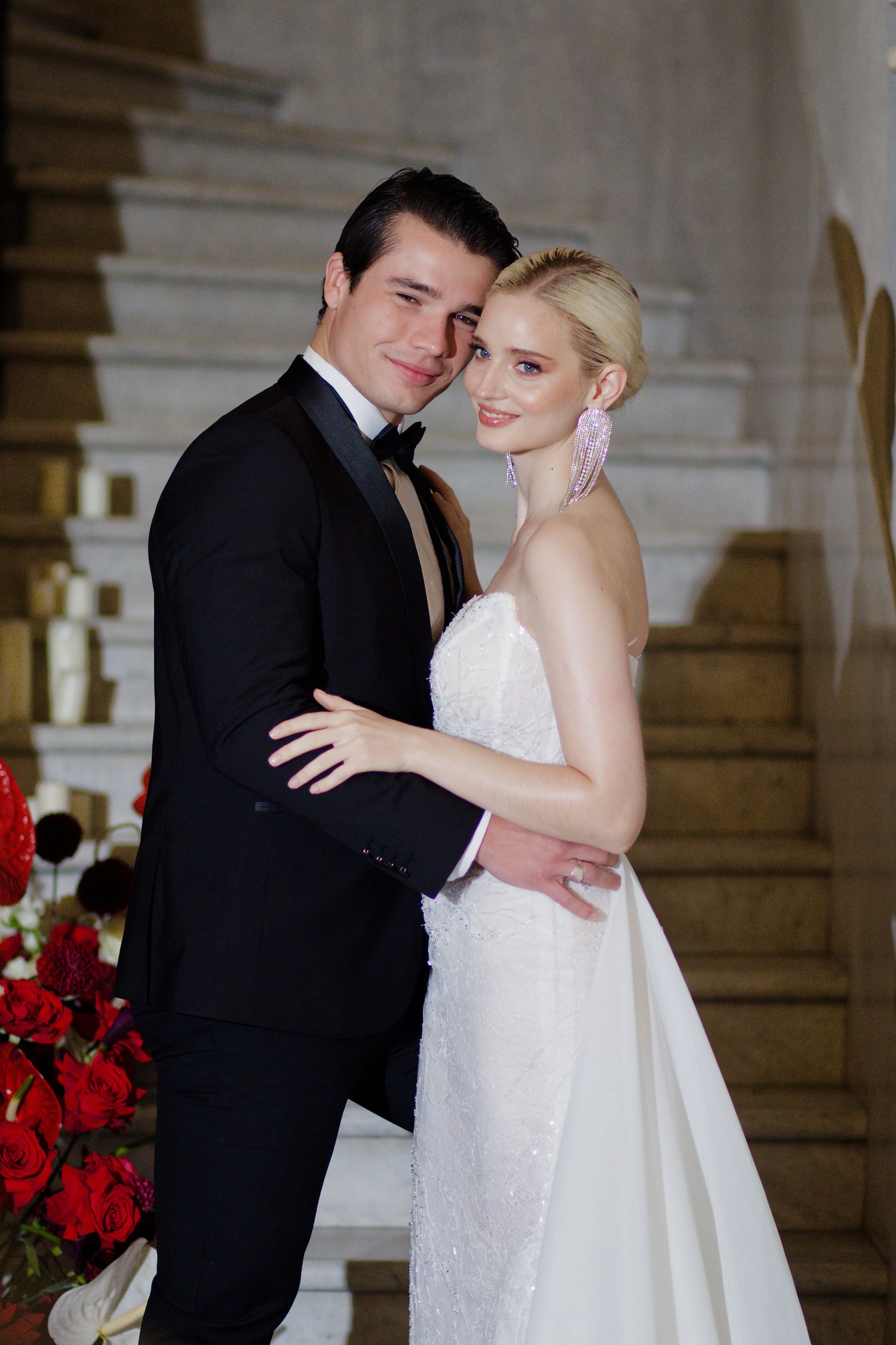 Romantic wedding couple embraces on ornate staircase with red floral decorations.