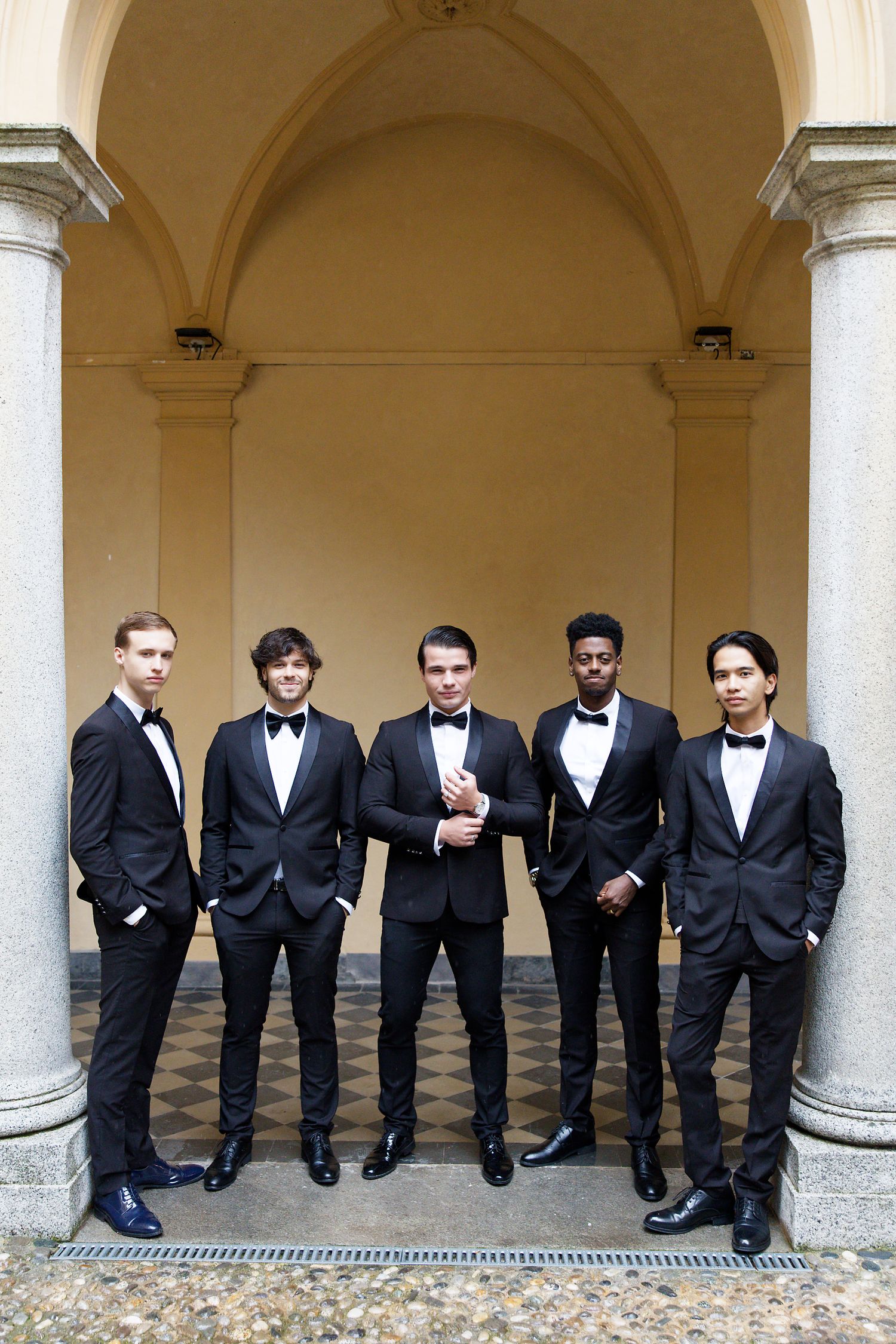 A group of five groomsmen in black tuxedos and bow ties pose together under an elegant arched colonnade.