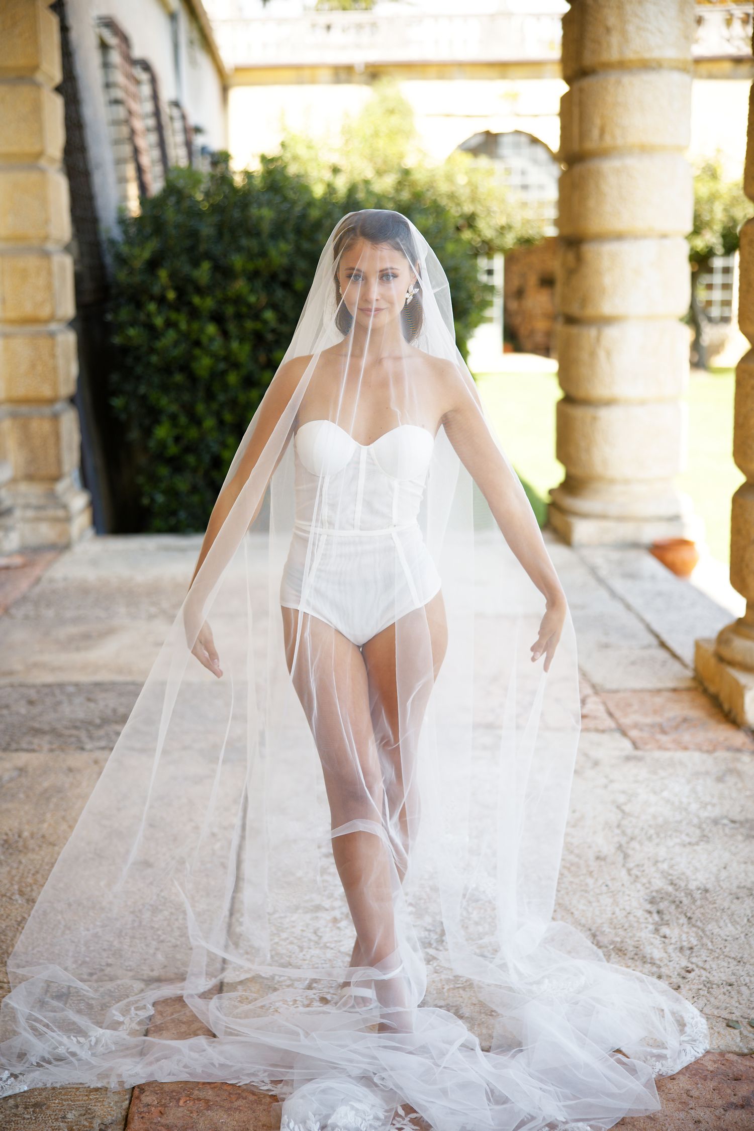 A bride wearing a white bodysuit and flowing cathedral veil poses in an elegant stone colonnade.