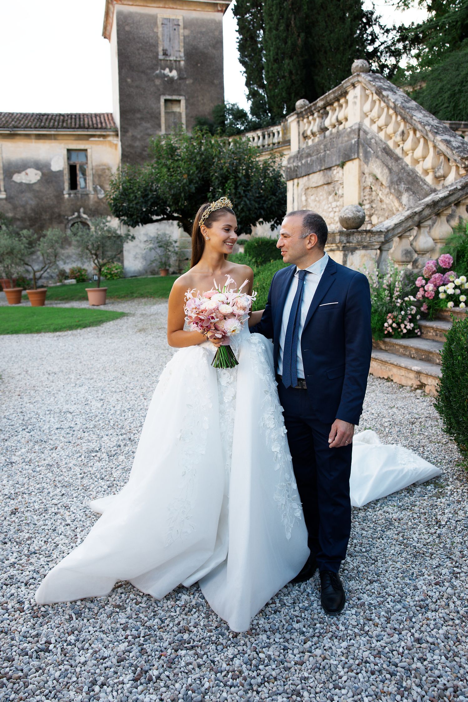 Wedding couple stands on a gravel path by ornate stone steps and garden at an elegant historic villa.
