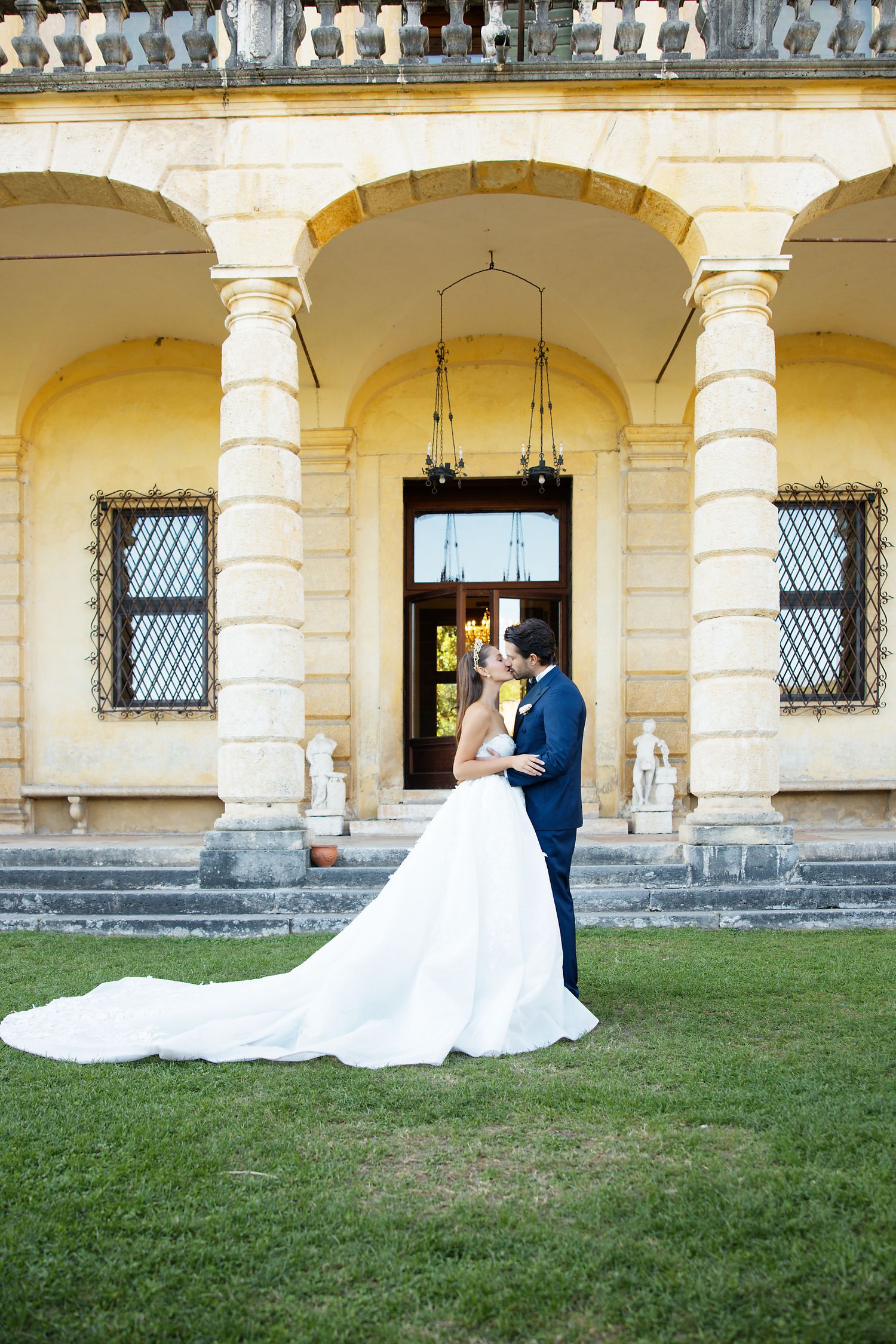 Wedding couple embracing on lawn in front of classical yellow villa with arched colonnade.