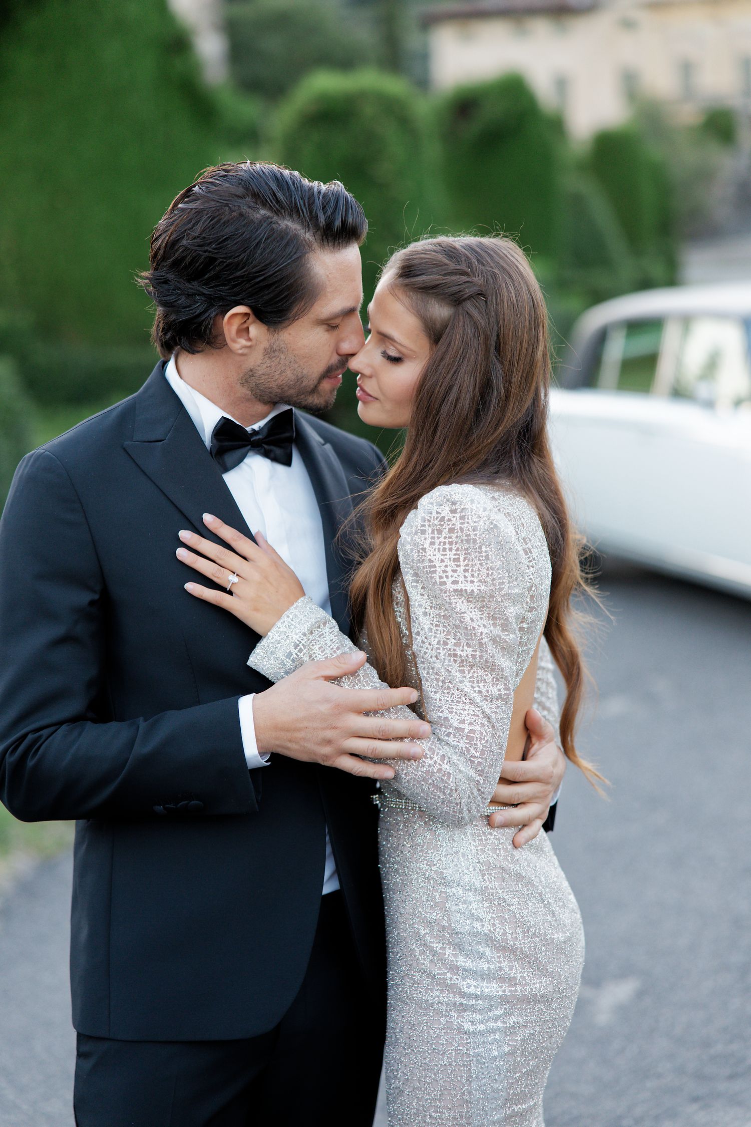 Couple in formal attire embrace intimately outdoors, bride wearing long sleeve sequined gown and groom in black tuxedo.