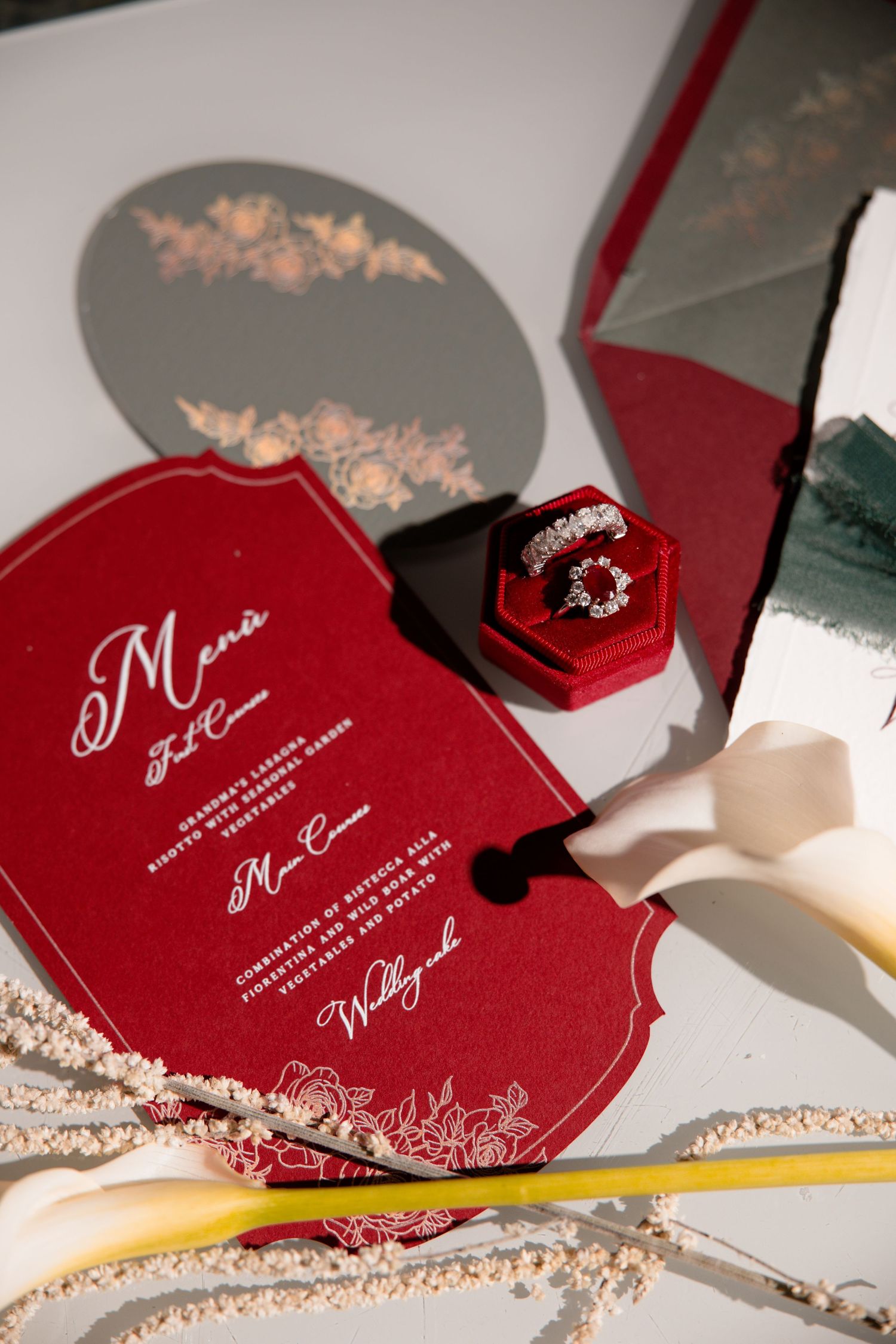 Red wedding invitation with decorative design and velvet ring box containing engagement rings on a white surface.