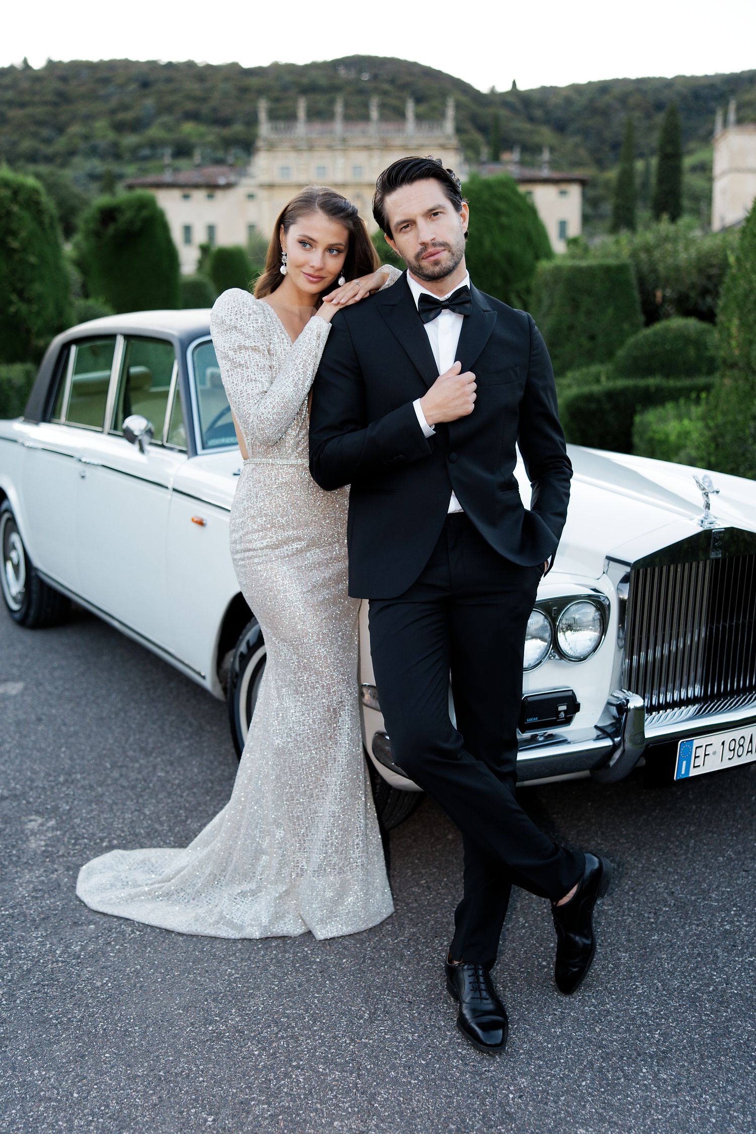 An elegant couple poses with a classic white Rolls Royce against an Italian villa backdrop.