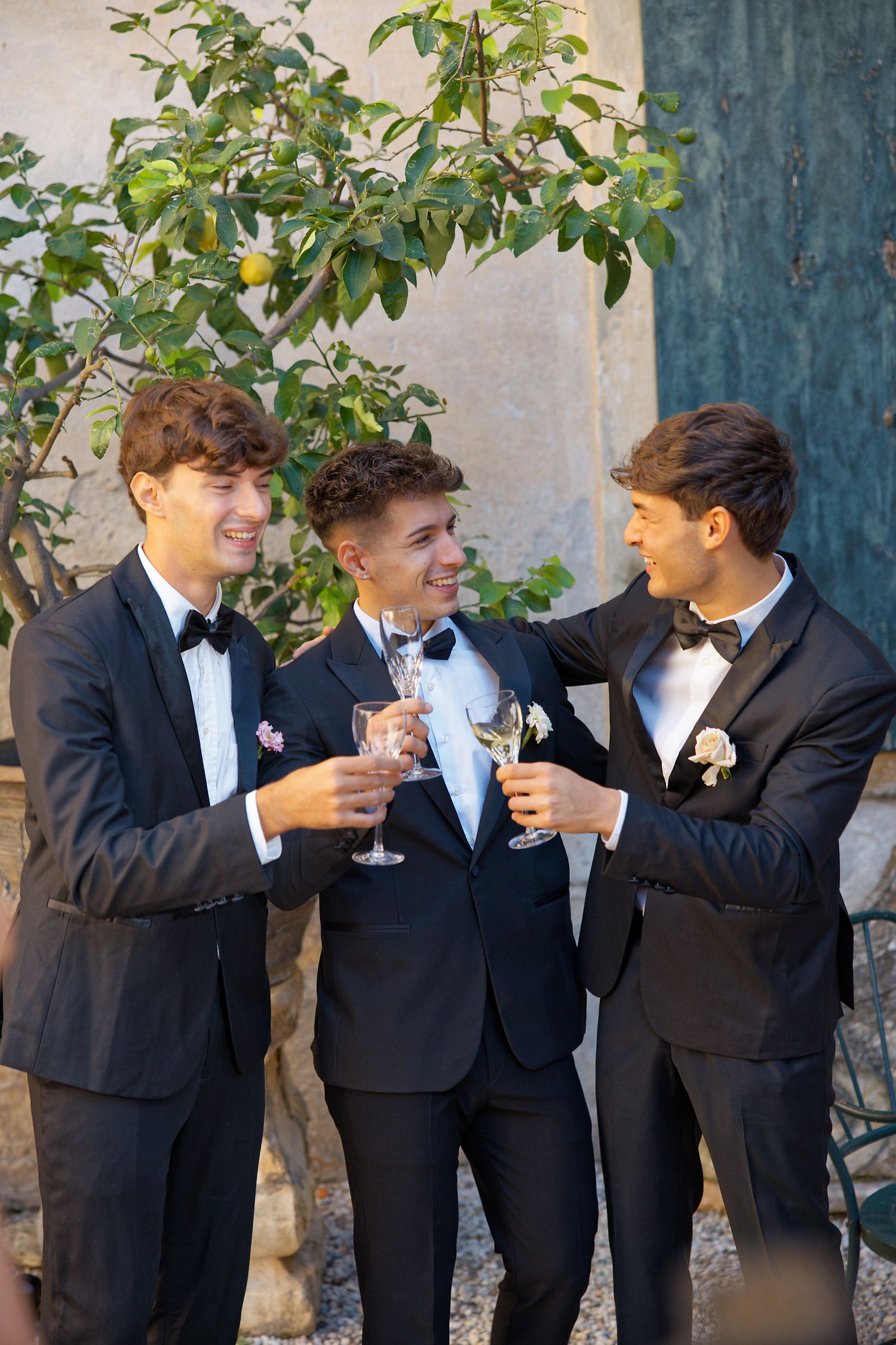 Three groomsmen in black tuxedos sharing drinks and laughing together outdoors.