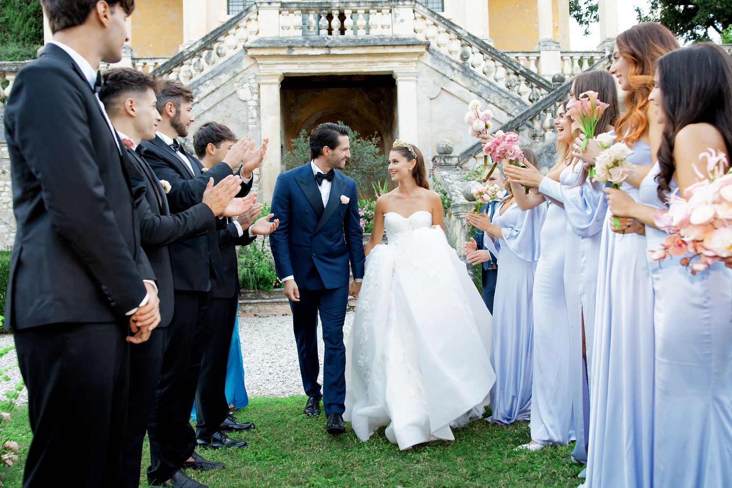 Bridesmaids in flowing blue gowns and groomsmen in dark suits celebrate outdoors at an elegant garden wedding ceremony.