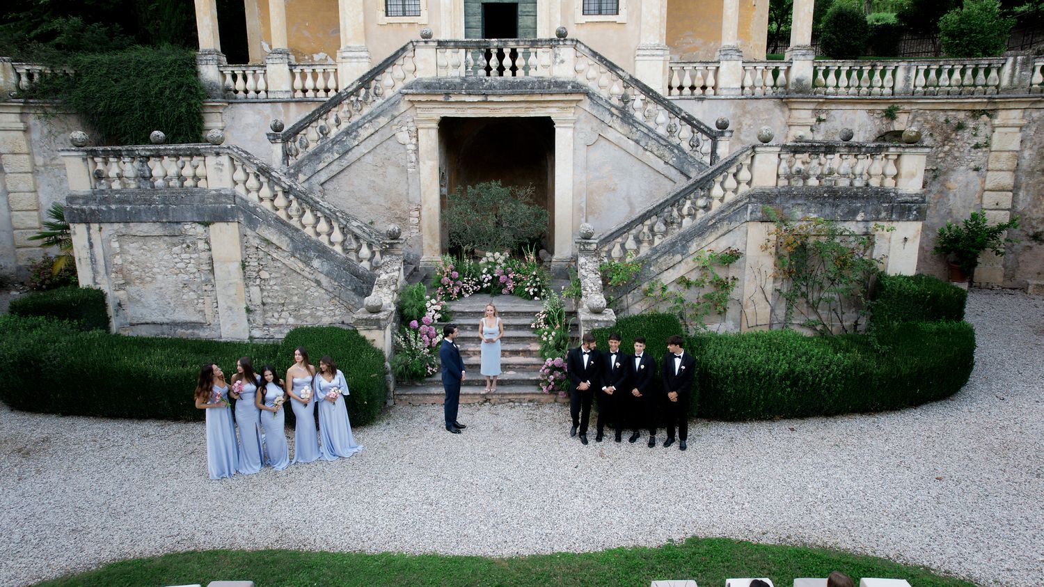 Aerial view of wedding party formation on symmetrical stone staircase with manicured hedges.
