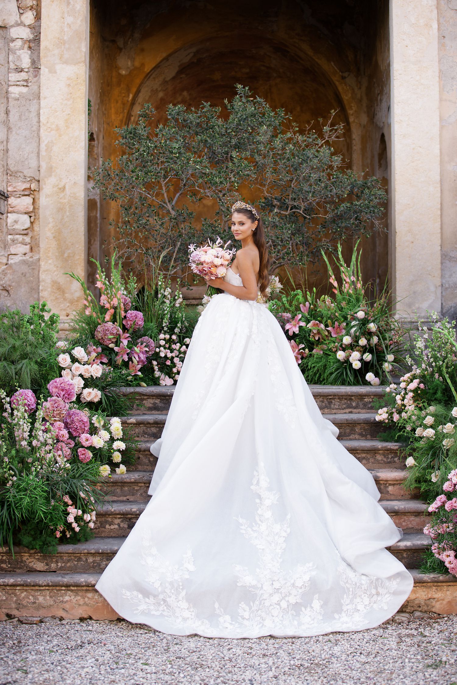 Elegant bridal portrait in flowing white gown against stone archway surrounded by pink and white garden flowers.