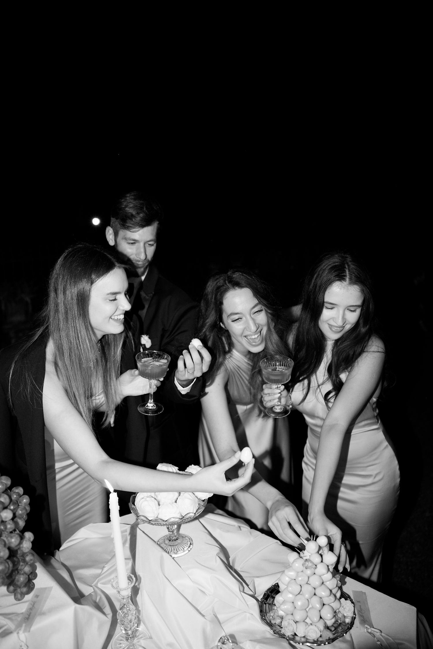 Black and white photo of wedding guests enjoying desserts and drinks at reception table.