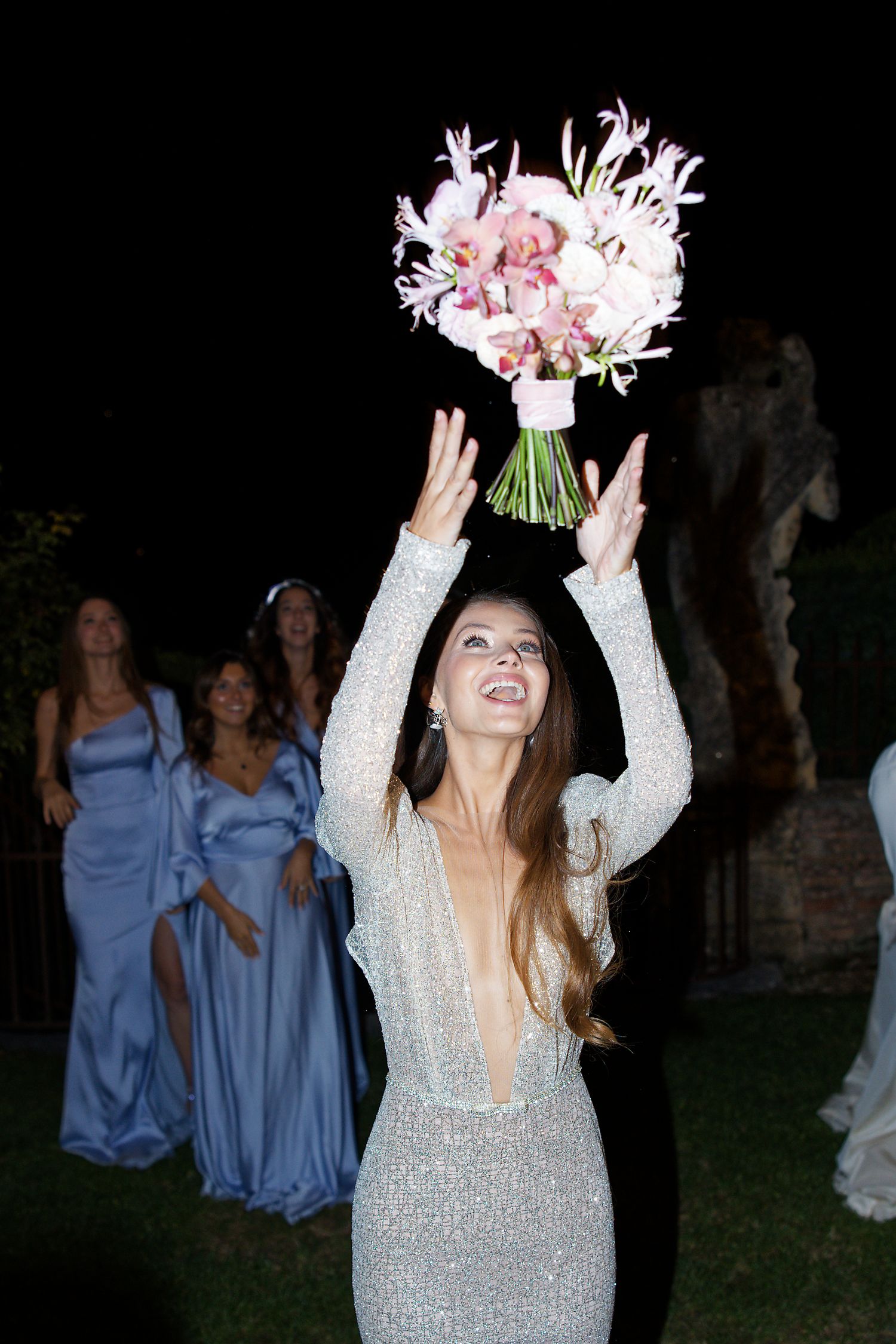 Bride in sparkly dress tosses bouquet while bridesmaids watch at nighttime wedding.