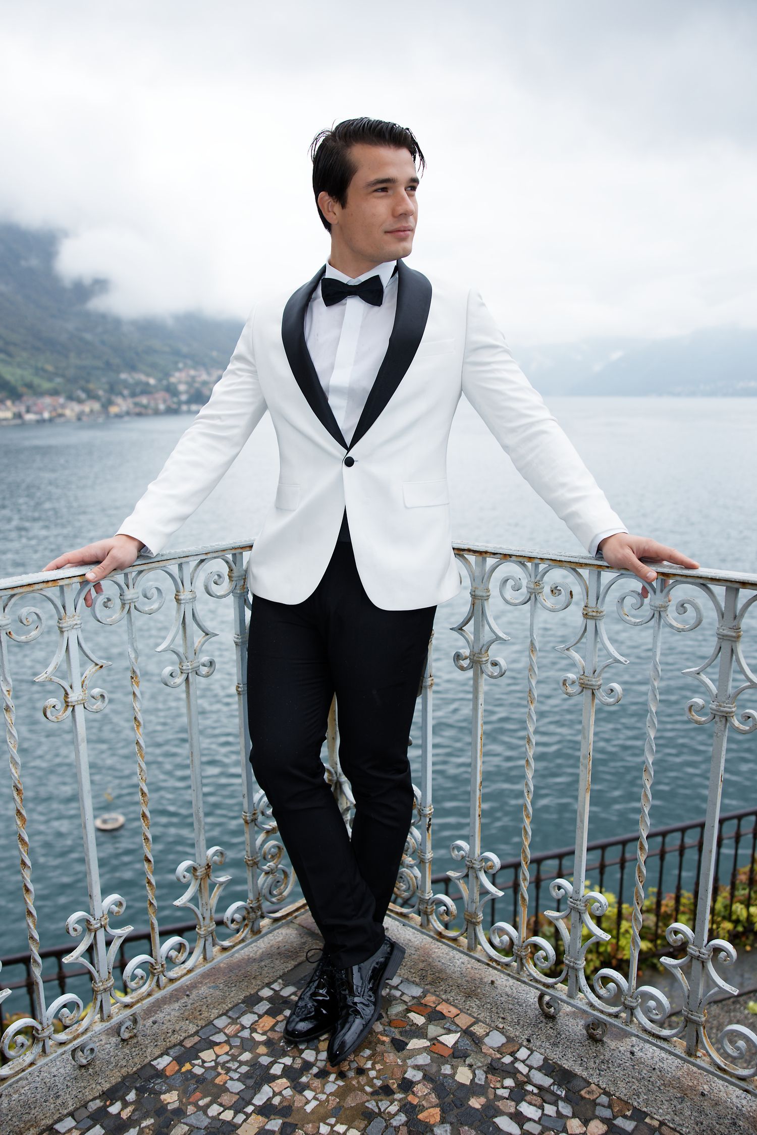 Groom in white tuxedo jacket poses on ornate balcony with lake and mountain backdrop.