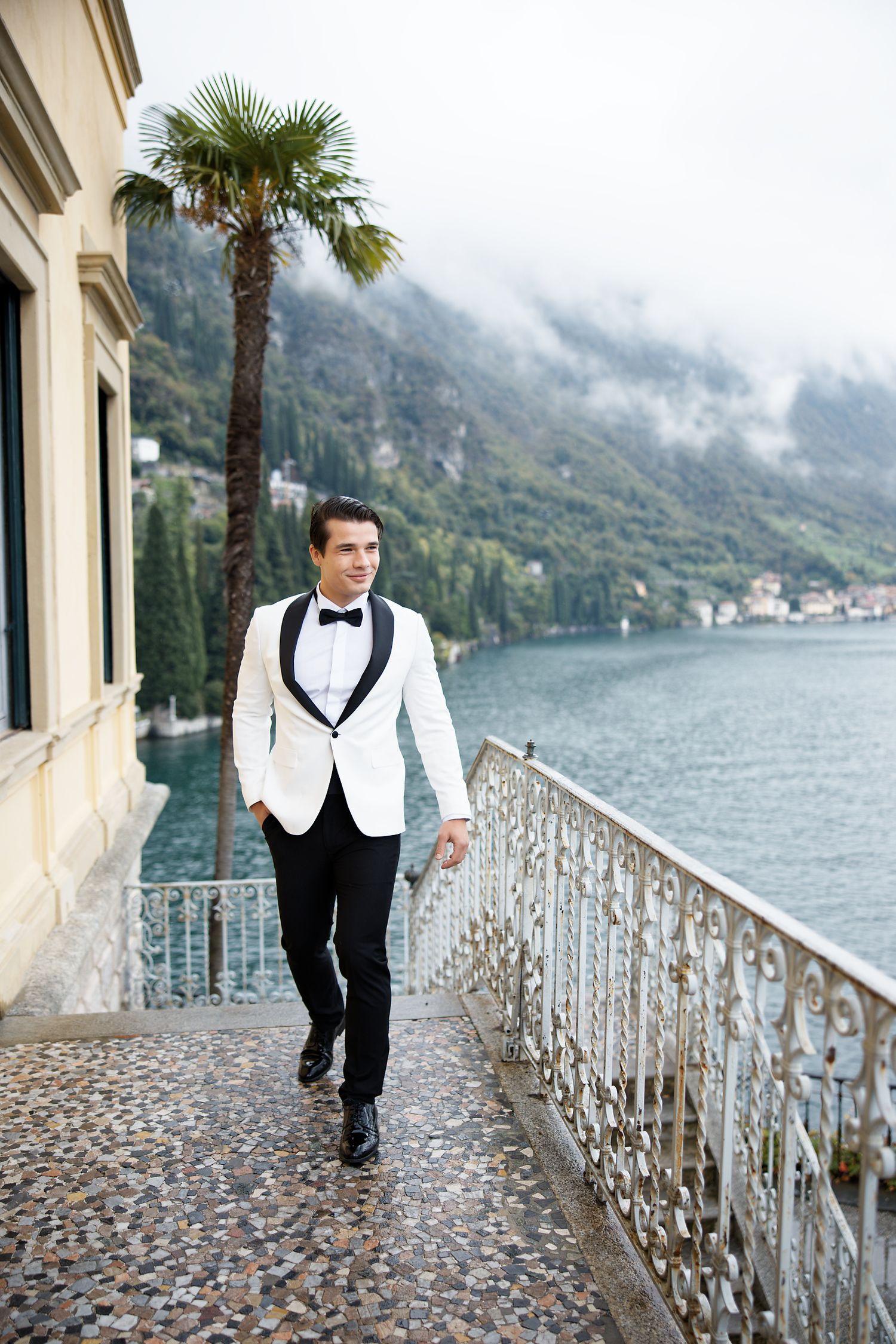 A groom in white tuxedo walks along a lakeside terrace with misty mountains in the background.
