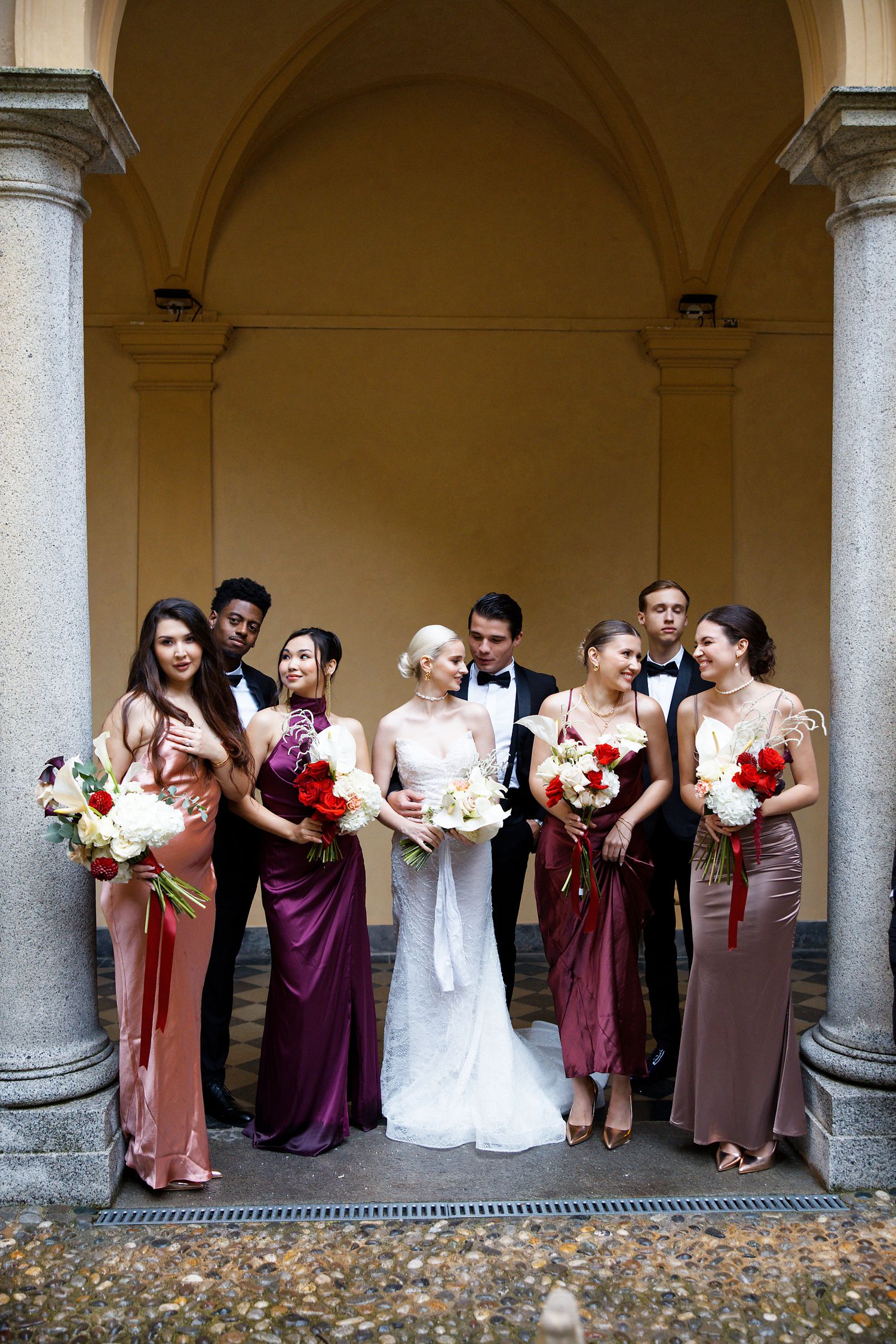 Wedding party posed under arched colonnade wearing elegant formal attire.
