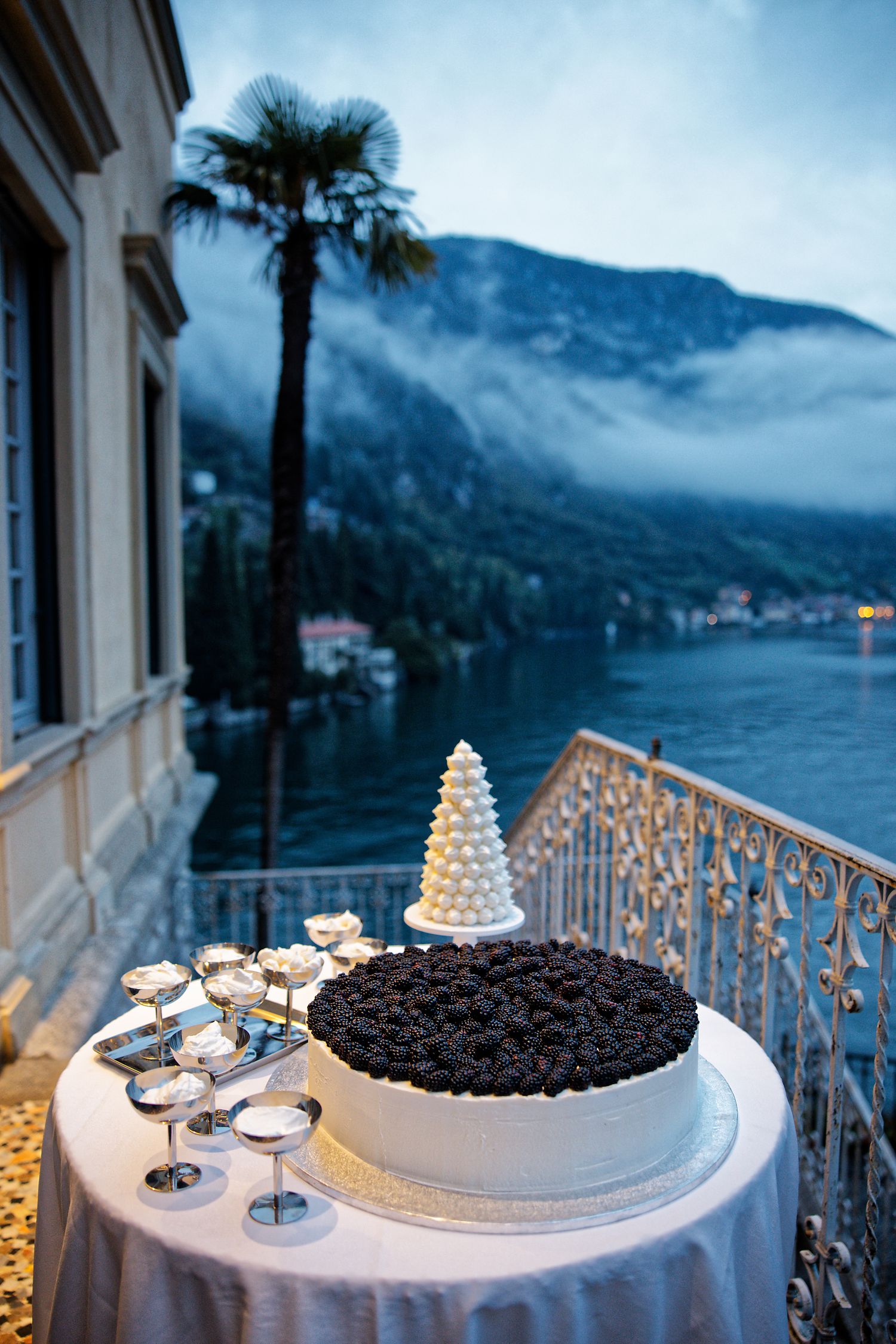Wedding dessert display on balcony overlooking Lake Como with mountains and palm trees at dusk.