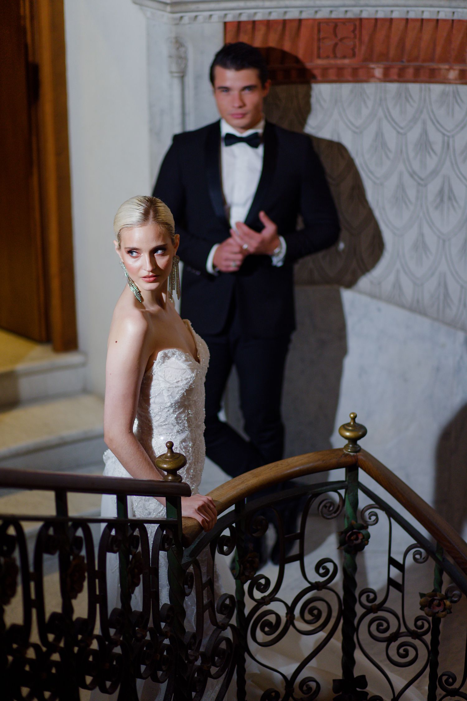 Elegant wedding couple posing on vintage staircase with ornate iron railings and decorative wall details.