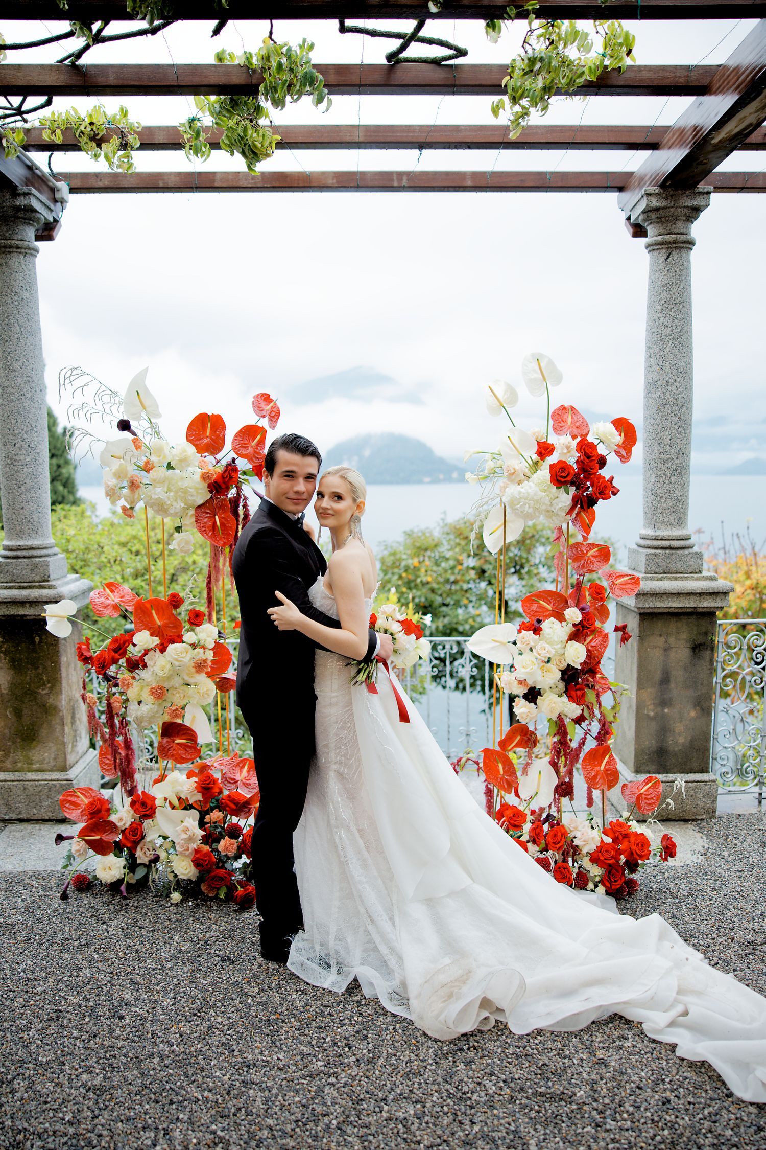 A wedding couple poses under a floral arch with orange and white blooms overlooking a lake.