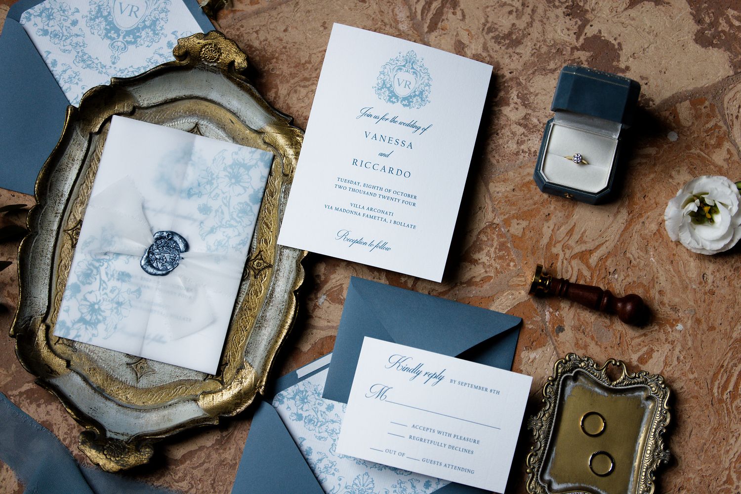 Elegant wedding invitation suite in blue and white with gold frames and ring box on textured surface.