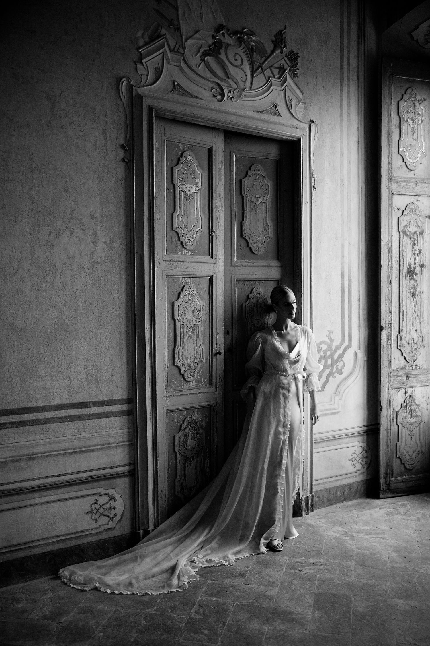 A dramatic black and white photo of a bride in a flowing dress standing by an ornate vintage doorway.