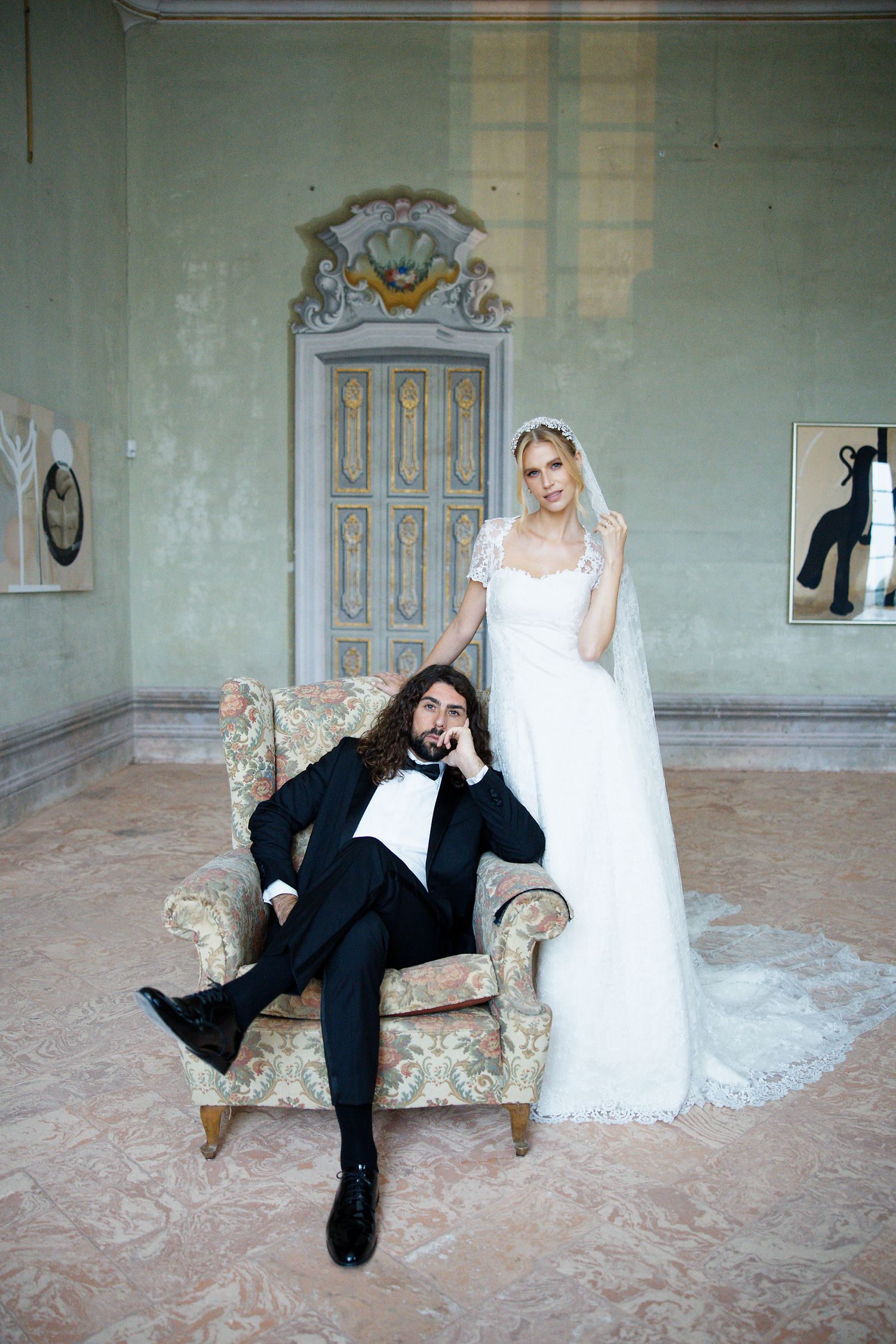 Whimsical wedding photo with groom lounging in vintage armchair while bride stands beside him.